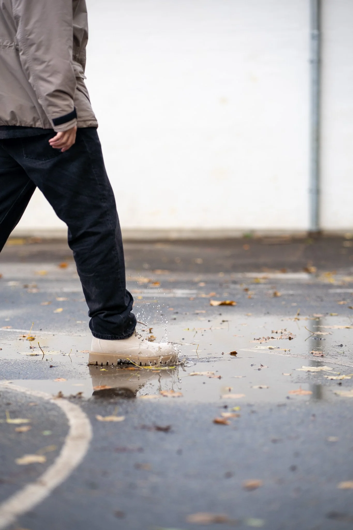 Person walking through a puddle on an outdoor court, wearing beige boots, black pants, and a beige jacket.
