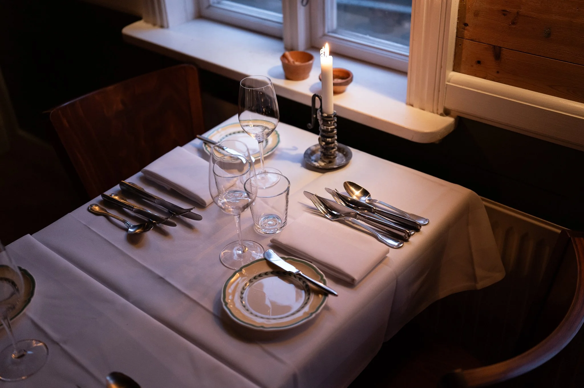A table set for dinner with a white tablecloth, multiple glasses, a plate with a patterned border, and silverware. A lit candle in a decorative holder is on the windowsill, with three small terracotta pots nearby in front of a wooden wall.