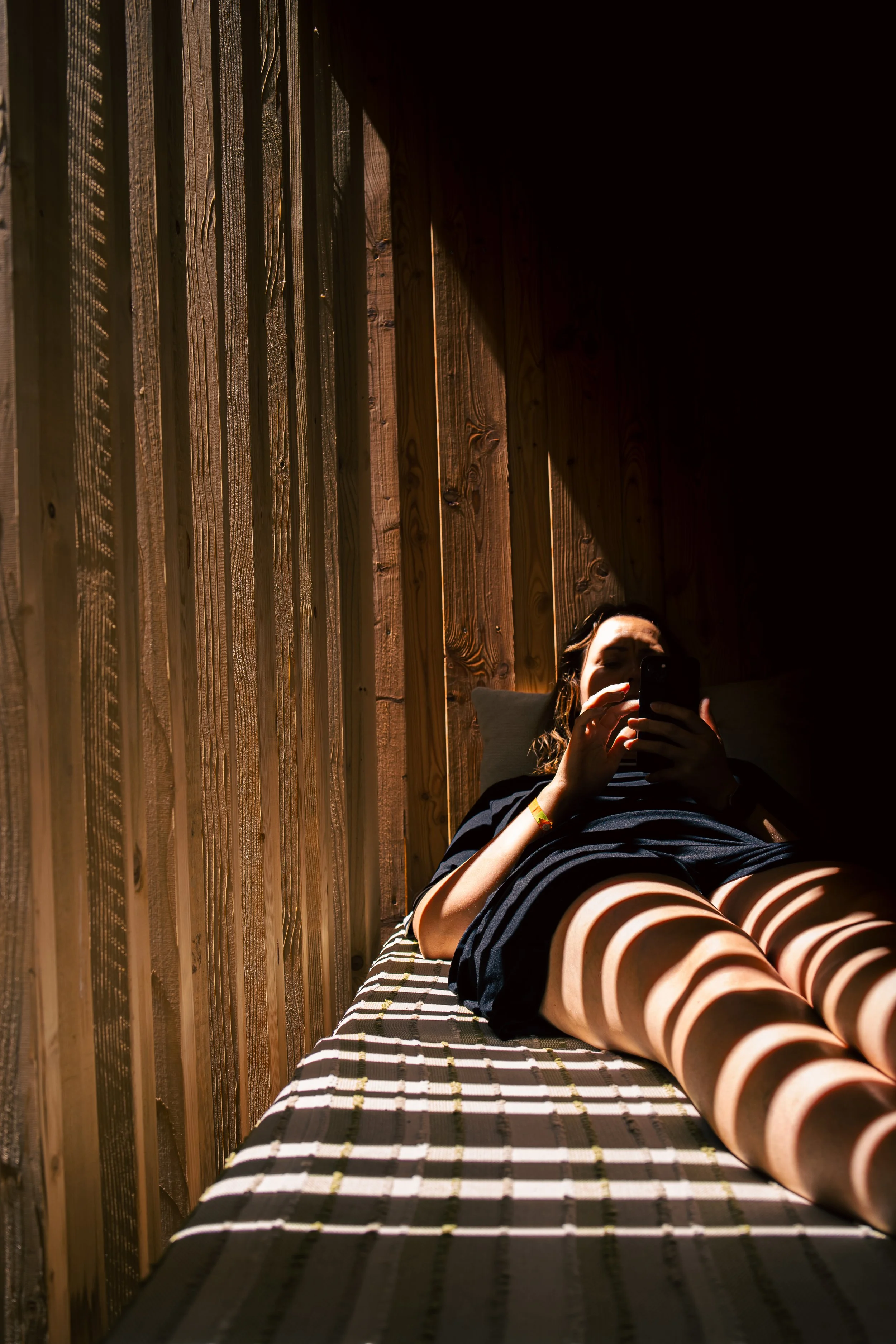 Relaxed woman lounging on a sunlit outdoor daybed, enjoying a quiet moment with her phone. Warm wooden textures and natural light create a calm, minimalist summer atmosphere.