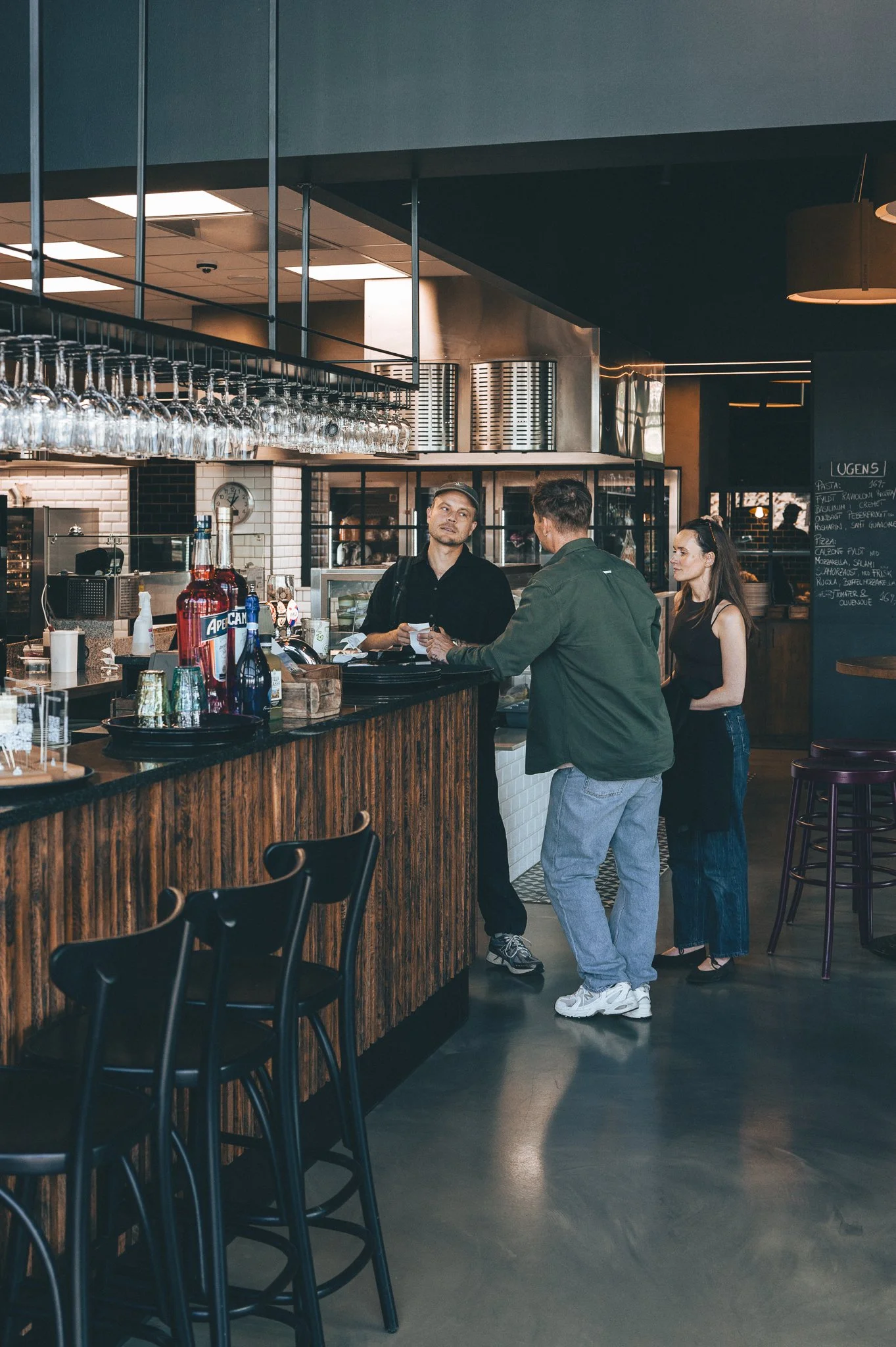 Three people are at a bar inside a restaurant. Two men are engaged in conversation at the counter, while a woman stands nearby observing. The bar has glasses hanging above, and a chalkboard menu is visible in the background.