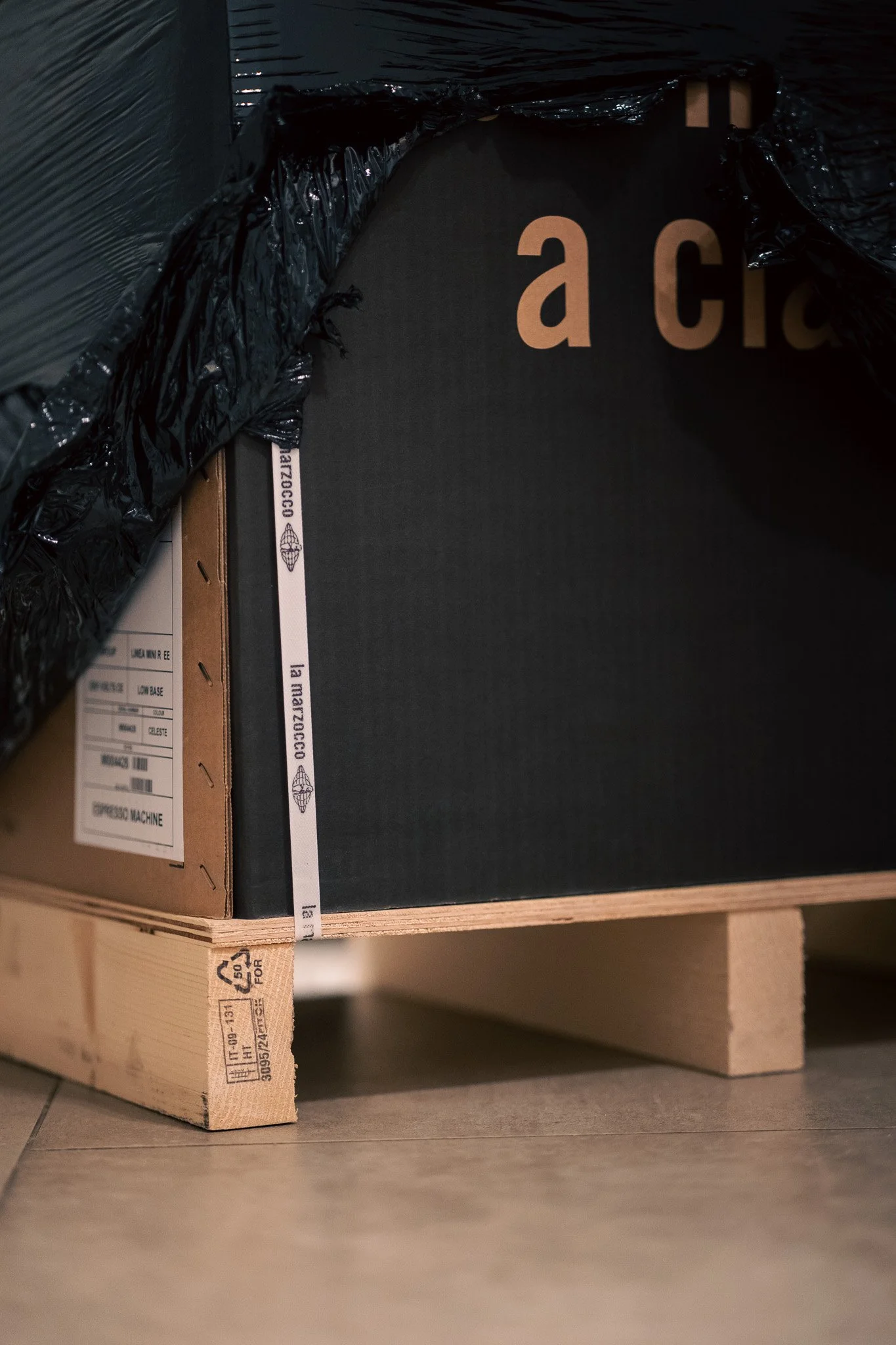 A black cardboard box partially covered by black plastic, placed on a wooden pallet, on a tiled floor.