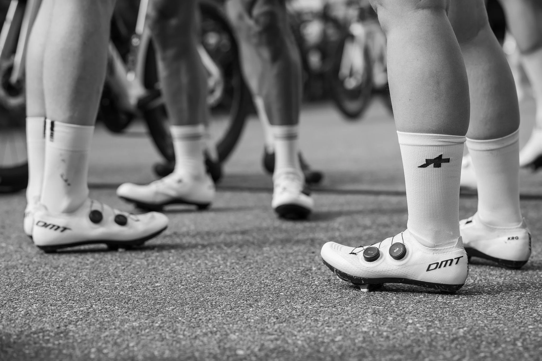 Close-up of cycling shoes and socks on the feet of cyclists in a lineup, with bikes in the background.
