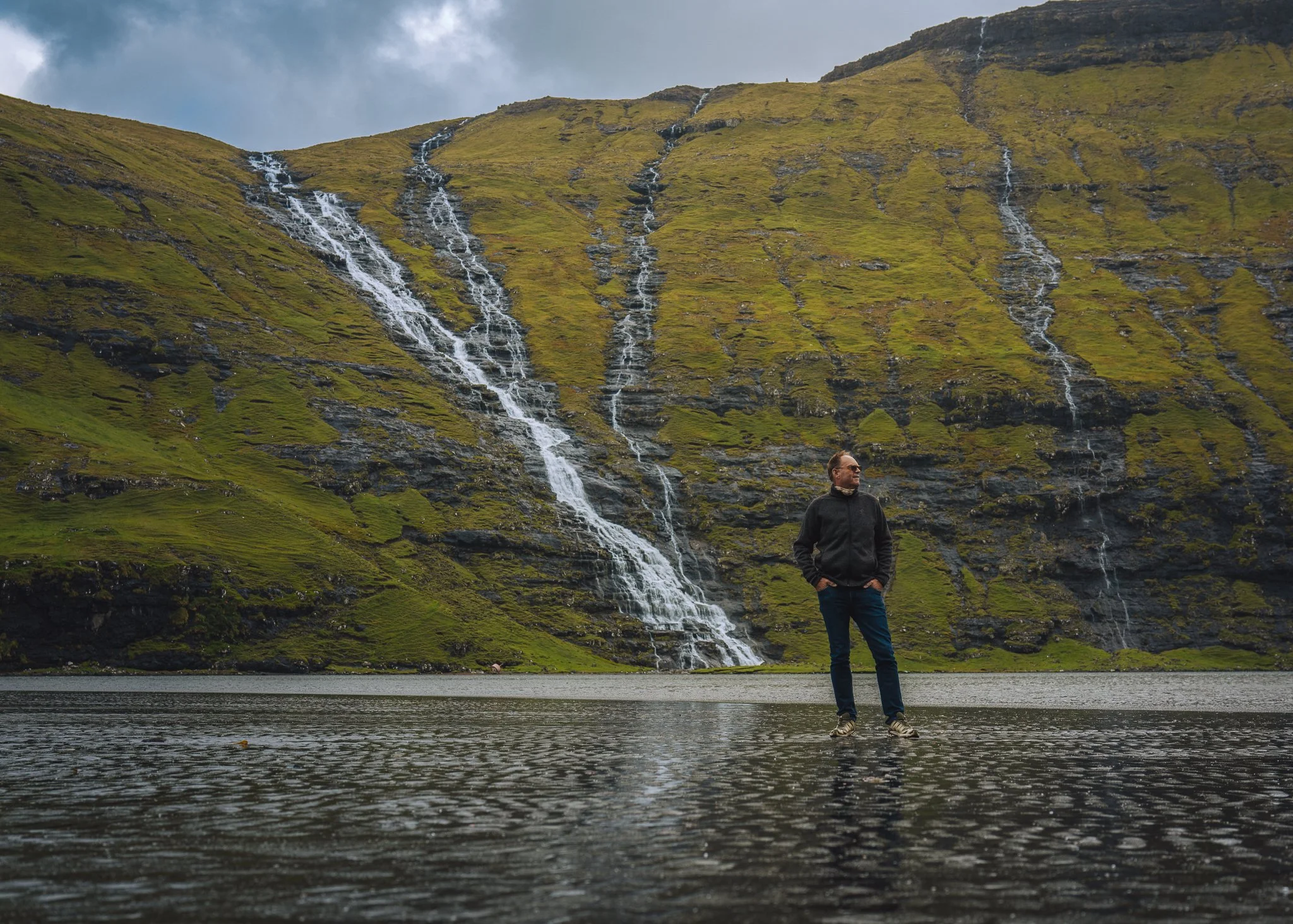 A man standing in shallow water in front of a large hillside with multiple waterfalls cascading down. The hillside is green with some dark rocks visible, and the sky above is cloudy.
