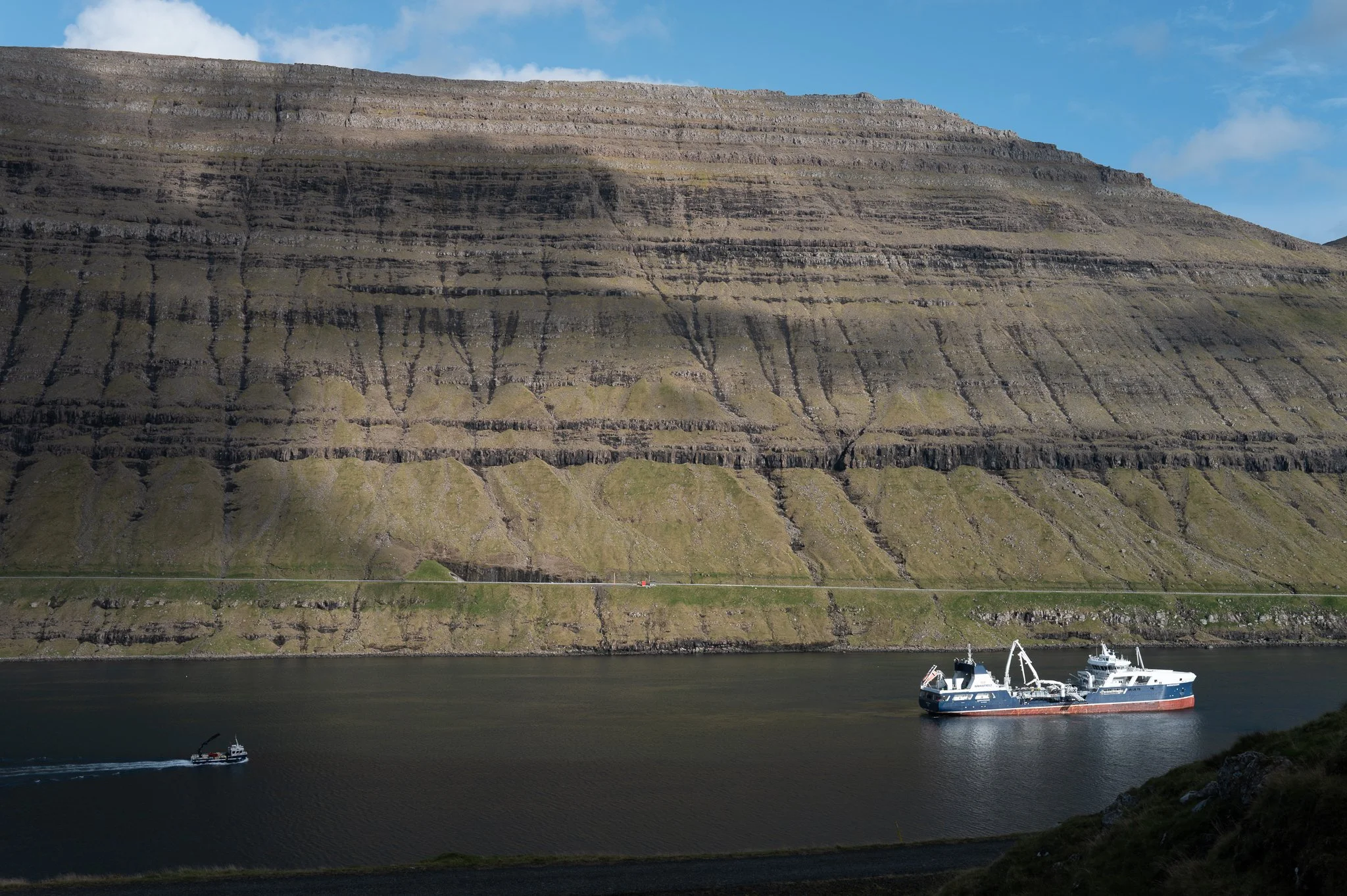 A large, layered cliffside with dark horizontal striations, next to water with a small boat and a larger ship, under a blue sky with scattered clouds.