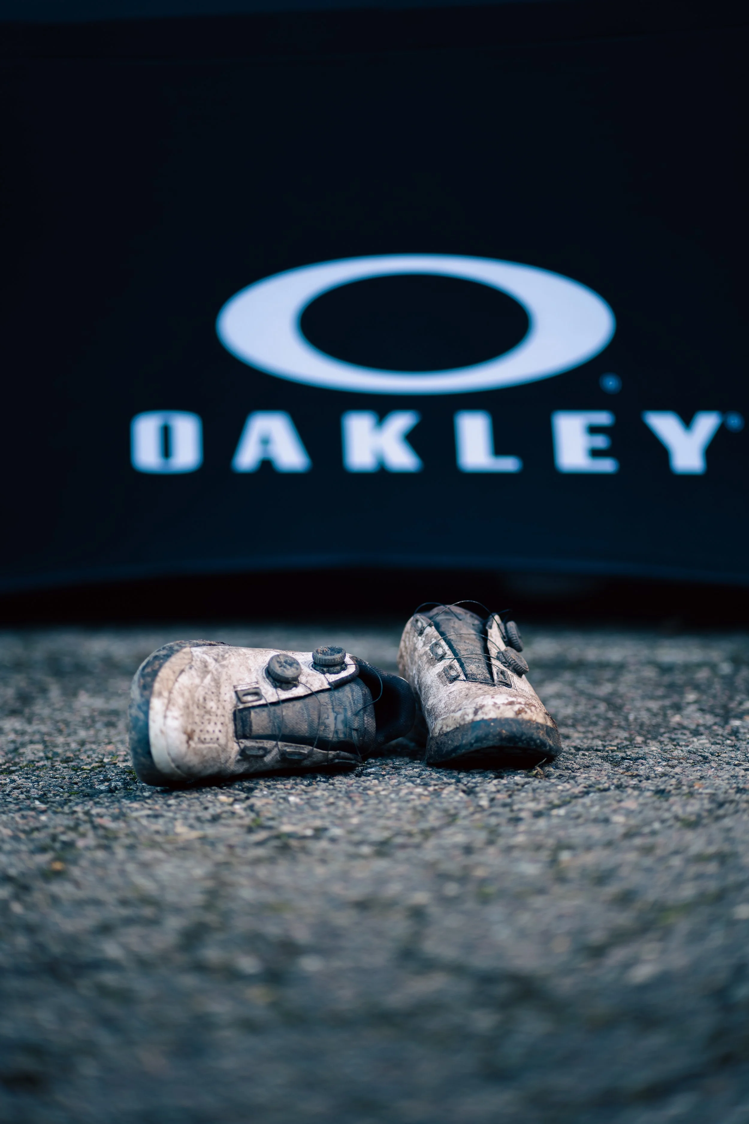 Worn cycling shoes resting on asphalt in front of a branded backdrop, symbolizing endurance, performance, and the raw reality of competitive cycling.