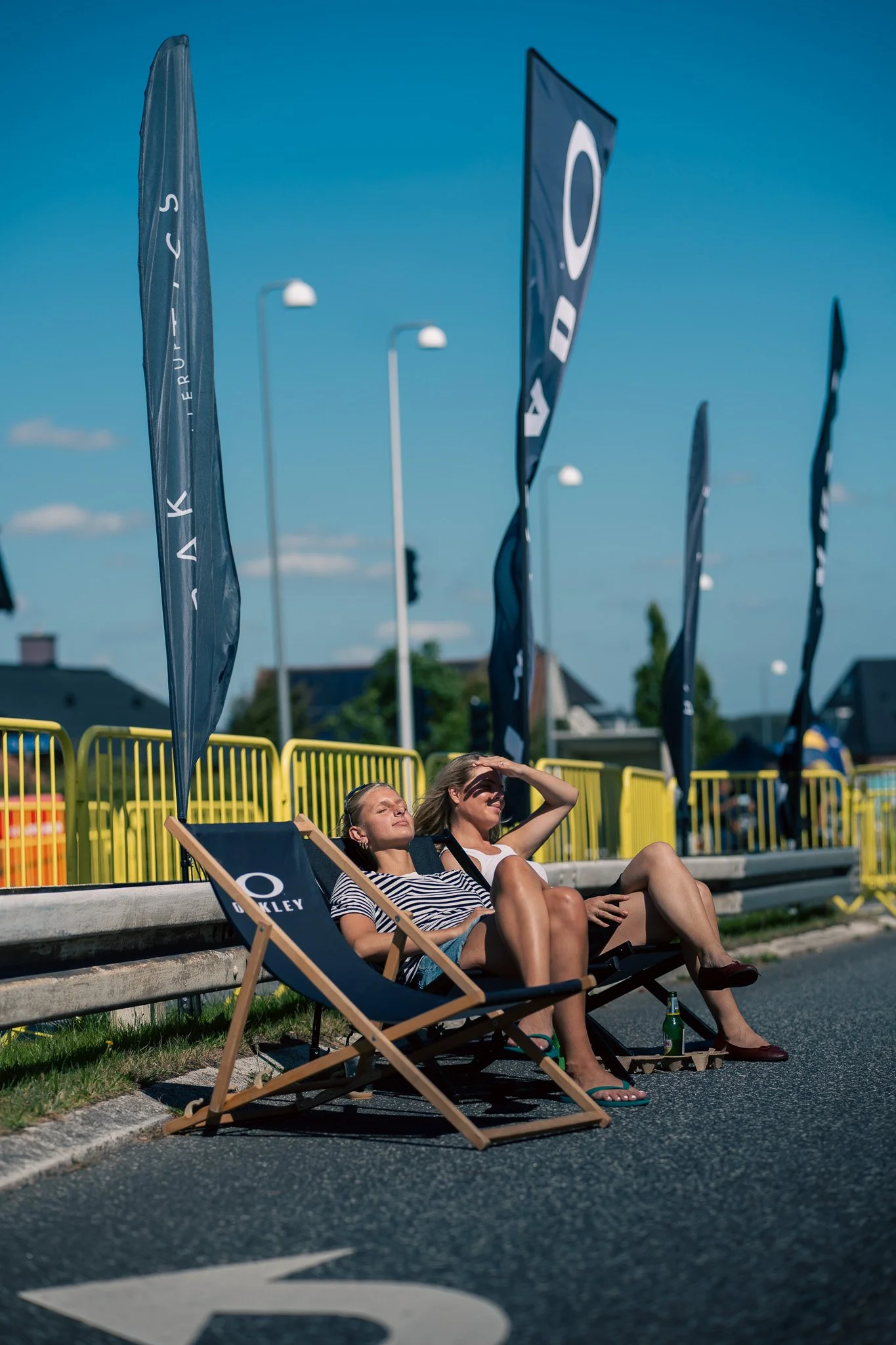 Two women relaxing on deck chairs on a roadside, shaded from the sun, during daytime, with flags and streetlights in the background.