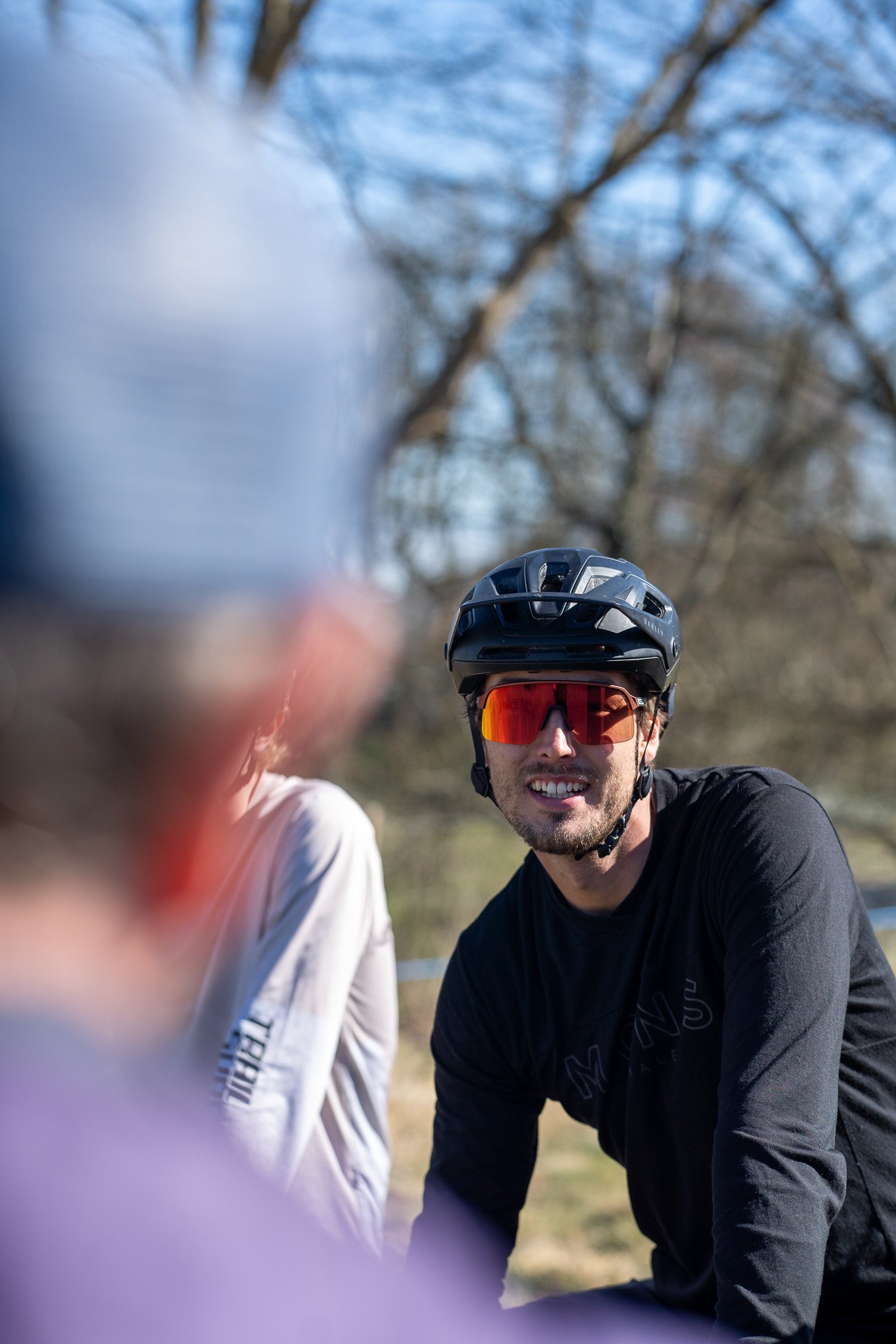 A man wearing a black helmet and orange sunglasses smiling outdoors with trees in the background.