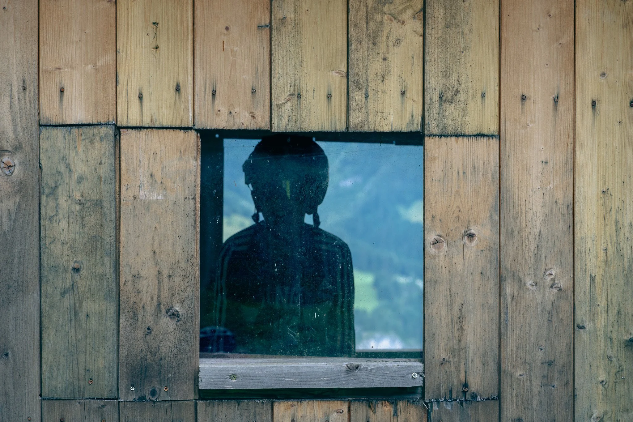 Silhouette of a person wearing a helmet, visible through a large glass window set in a wooden wall with weathered vertical planks.