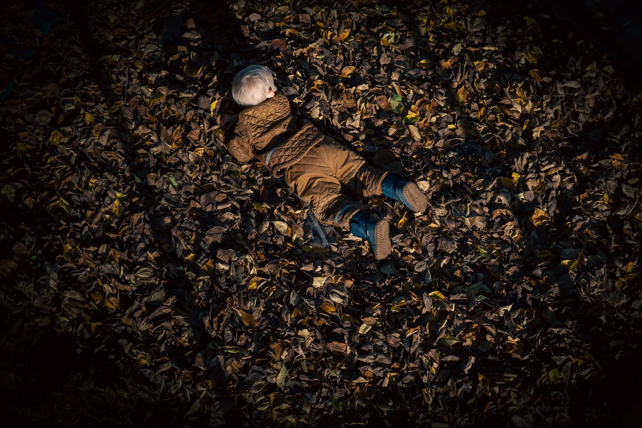 A young child with light-colored hair is lying on their stomach in a bed of fallen autumn leaves, wearing a brown quilted jacket, brown pants, and dark shoes.
