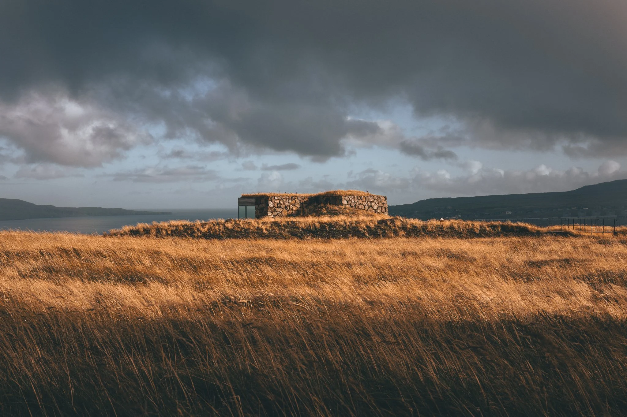 A stone building with a grass-covered roof on a hill, overlooking a body of water, under a cloudy sky.