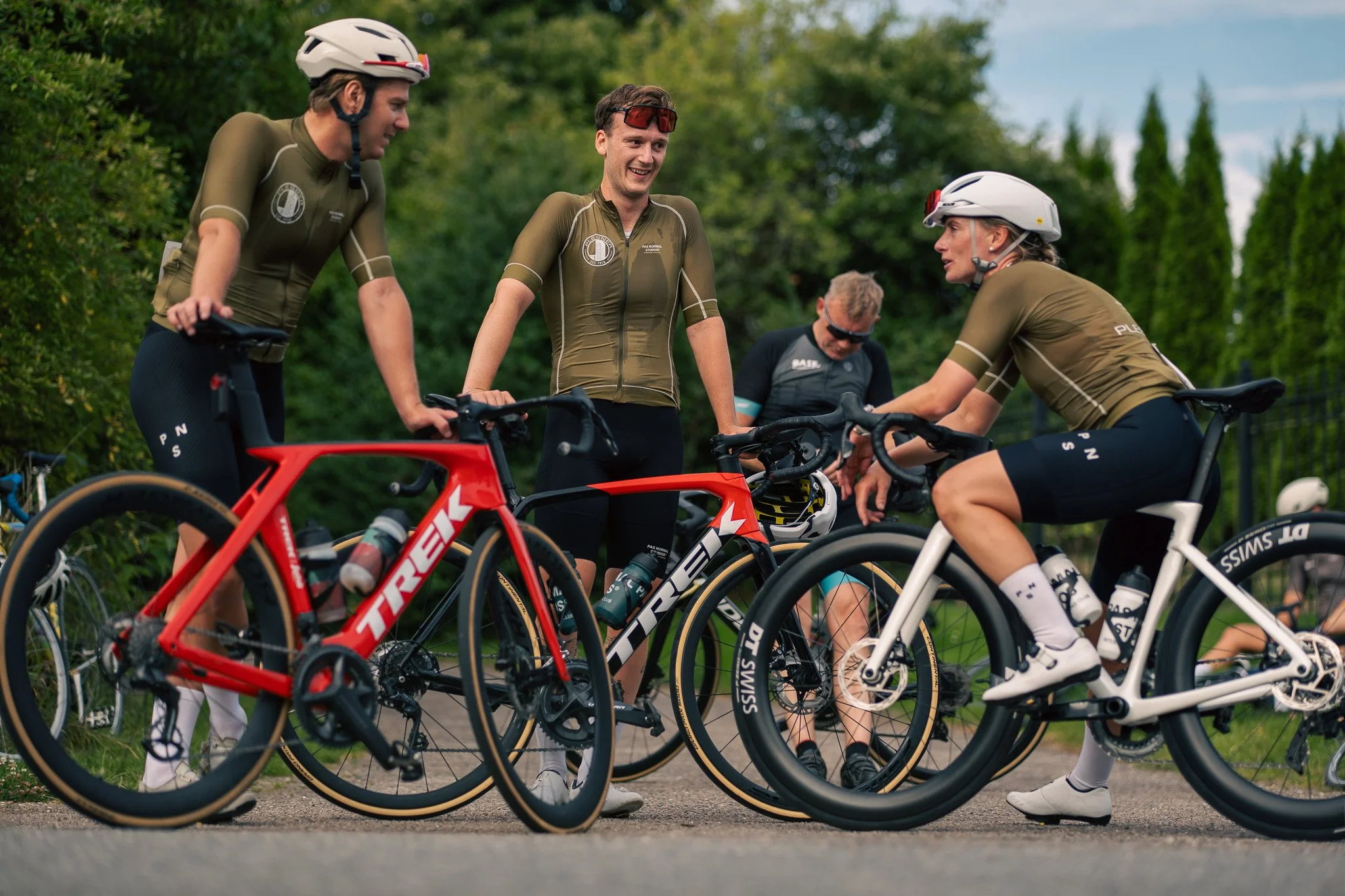 Four cyclists in workout gear have a conversation beside their bikes outdoors, with trees in the background.