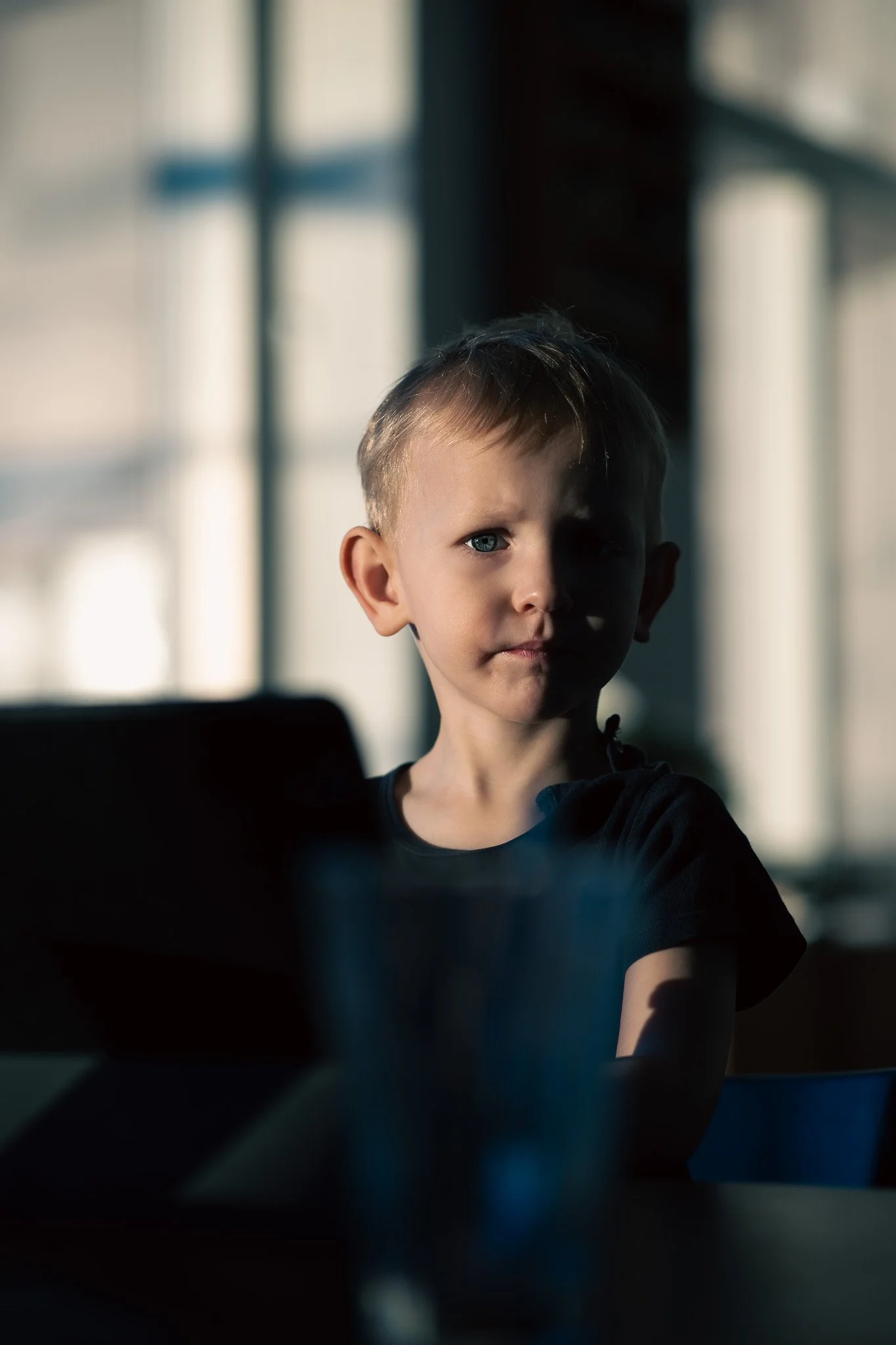 A young boy with light hair and blue eyes sits indoors in shadow, with sunlight streaming through large windows behind him.