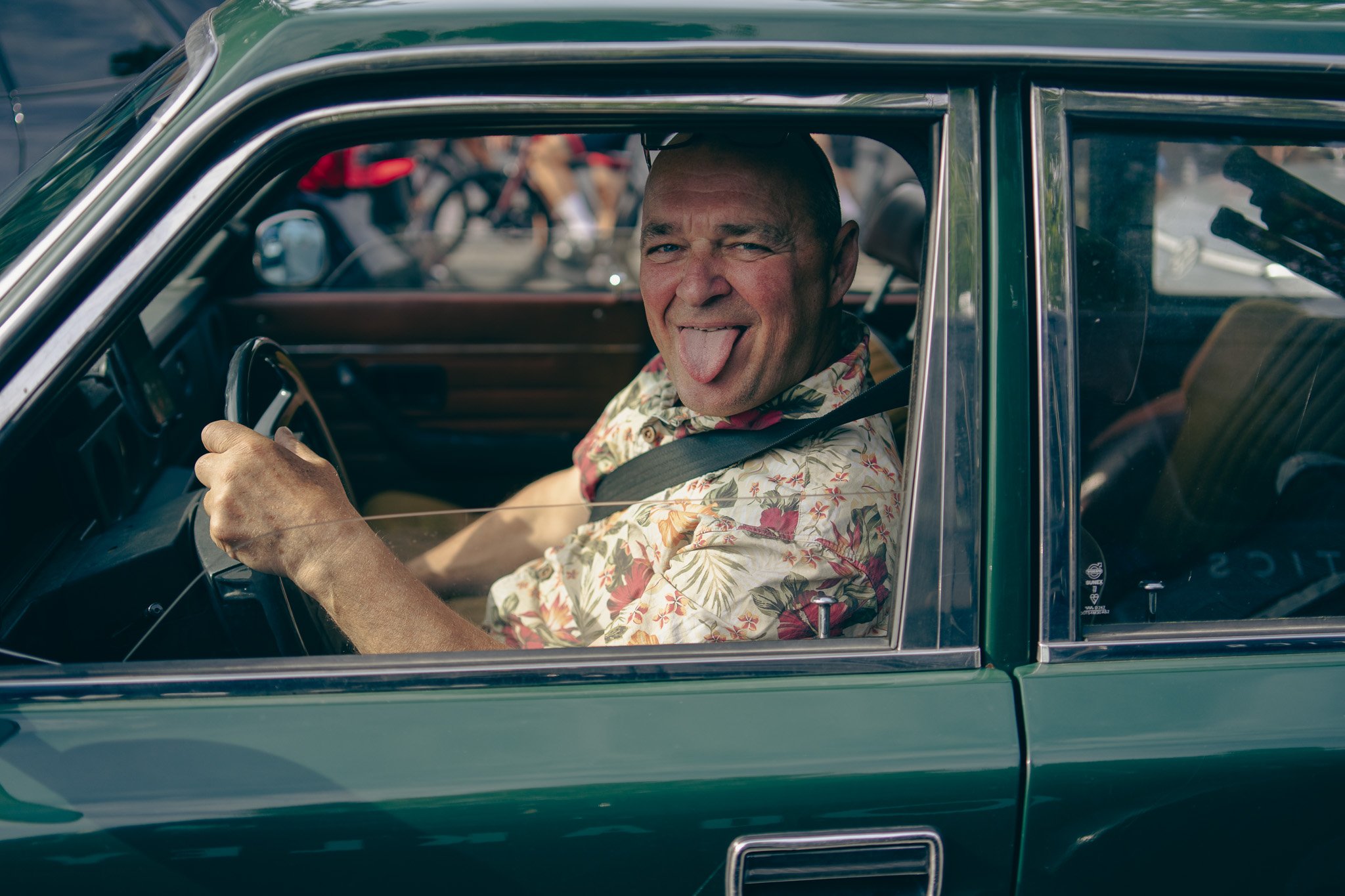 A man in a floral shirt sitting in a green vintage car, sticking out his tongue and smiling.