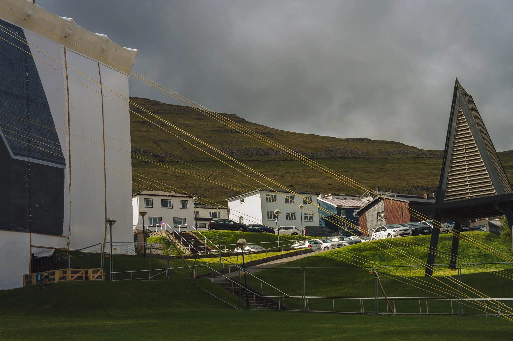 Residential area with parked cars, power lines, and modern houses on a hillside under cloudy sky.