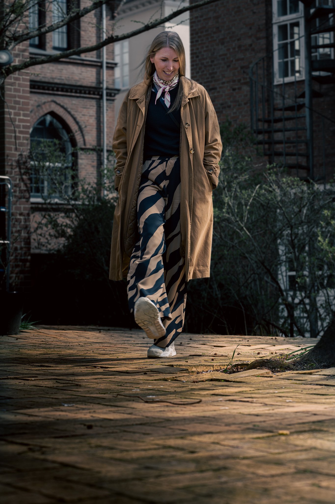 A woman with blonde hair walking on a brick pathway outdoors in an urban residential area, wearing a brown trench coat, black top, patterned wide-leg pants, and white sneakers.