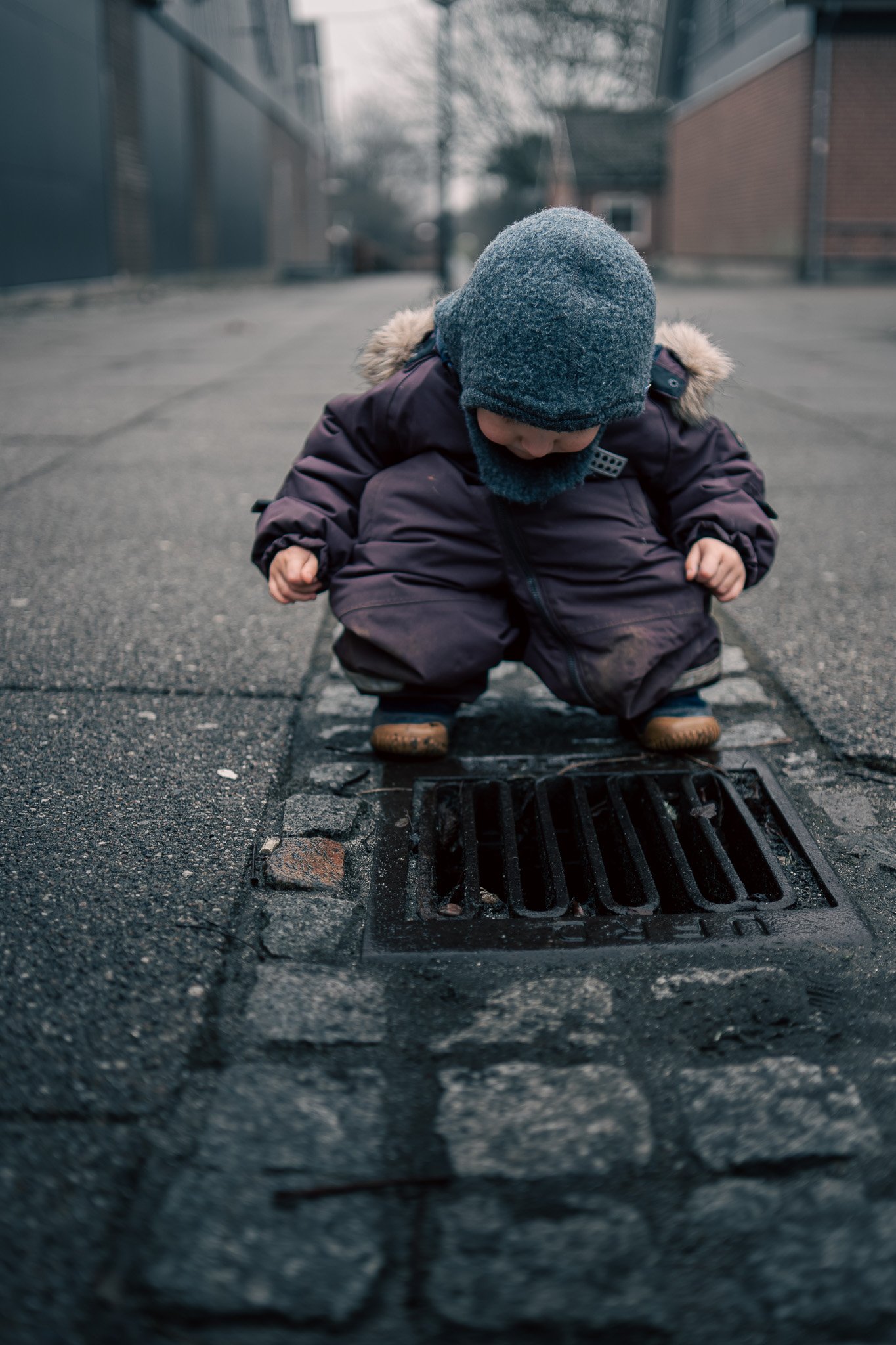 A young child in a winter coat and hat squatting down on a sidewalk, examining a street drain grate.