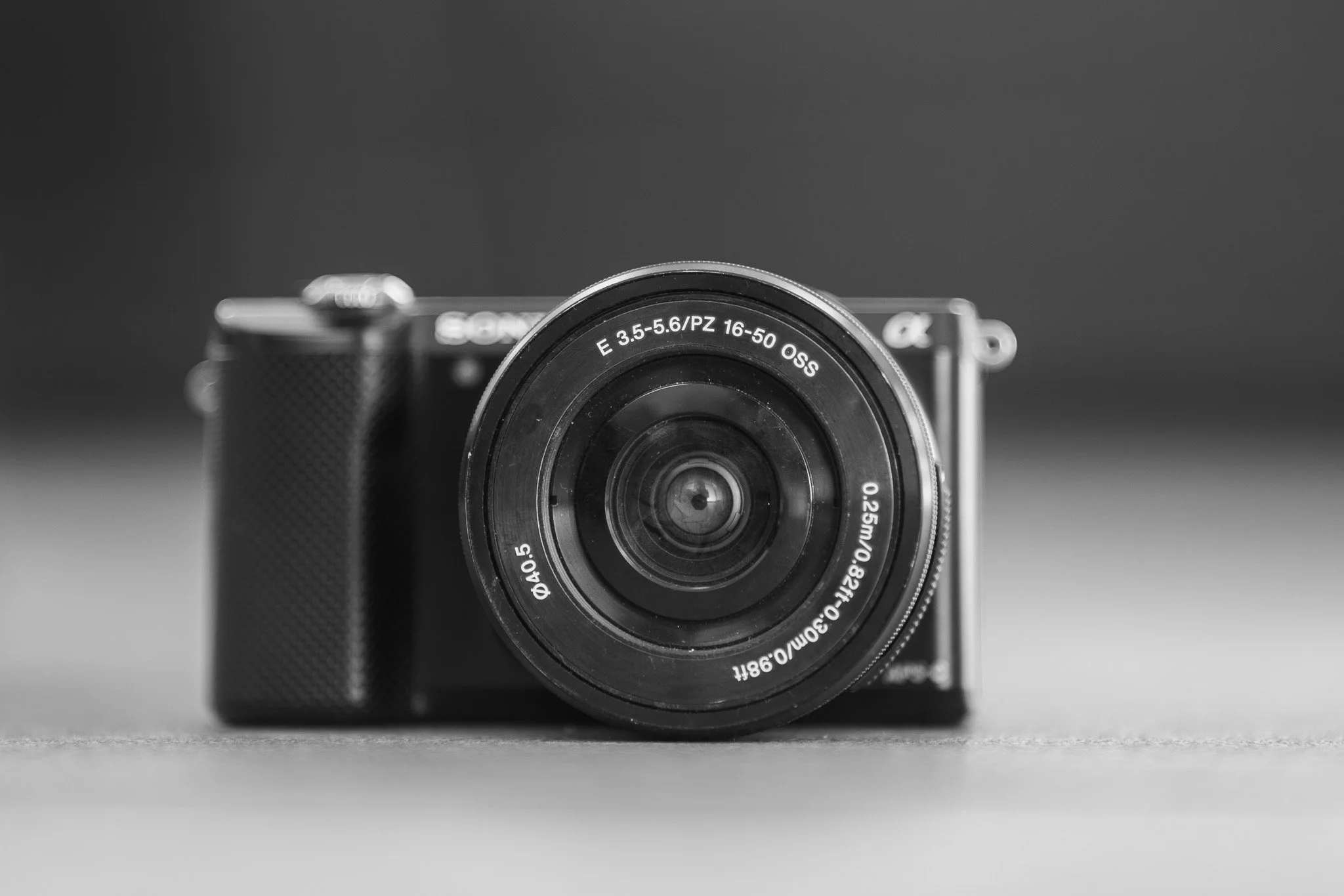 Black and white photo of a Sony mirrorless camera with a zoom lens, placed on a flat surface against a plain background.