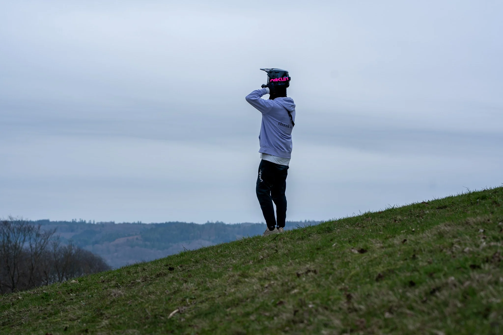 Person standing on a grassy hillside, wearing a helmet and hoodie, looking at the horizon during overcast weather.