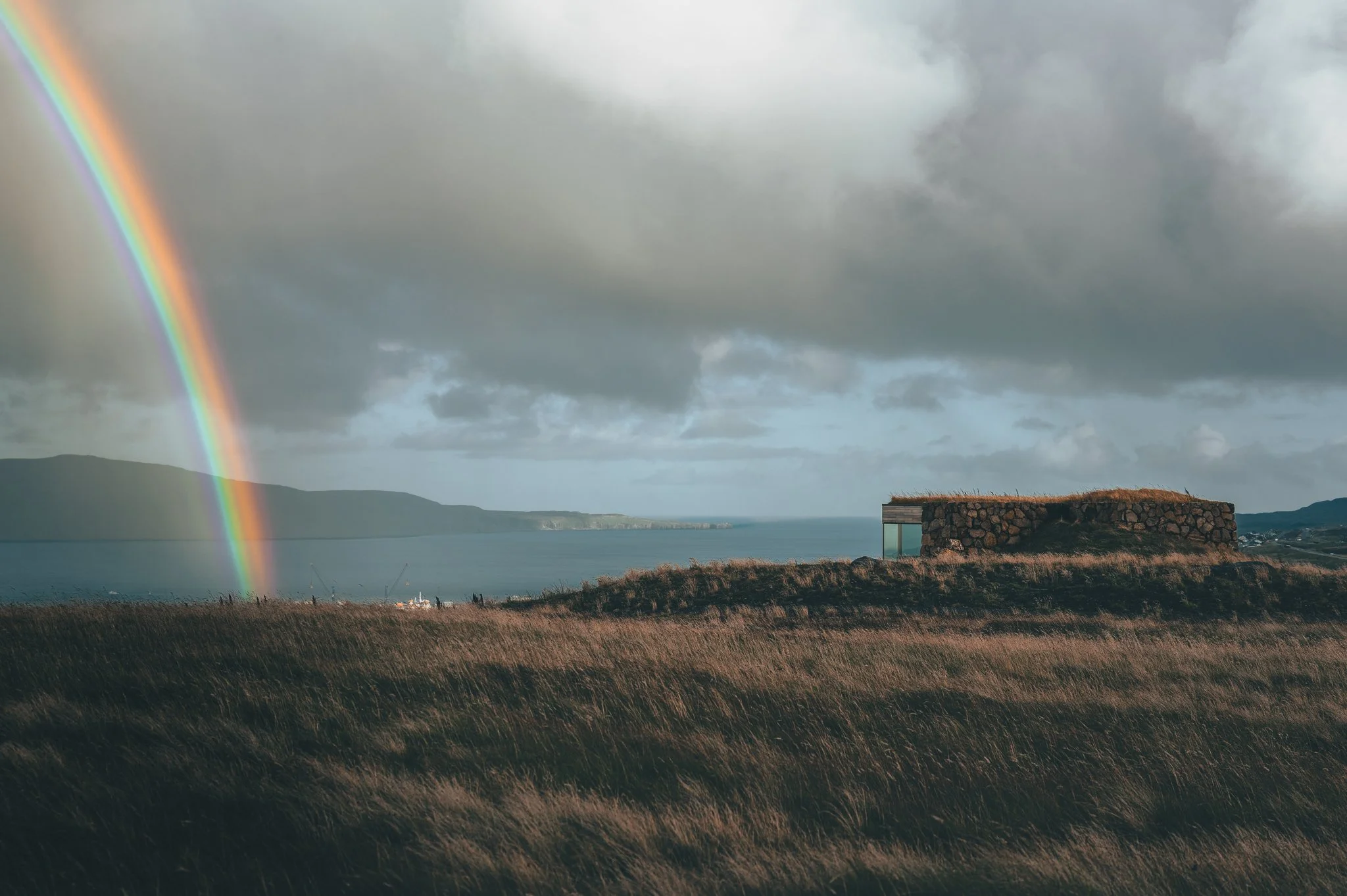 A rainbow arcs across a cloudy sky over a coastal landscape with grassy fields, water, and hills in the background.
