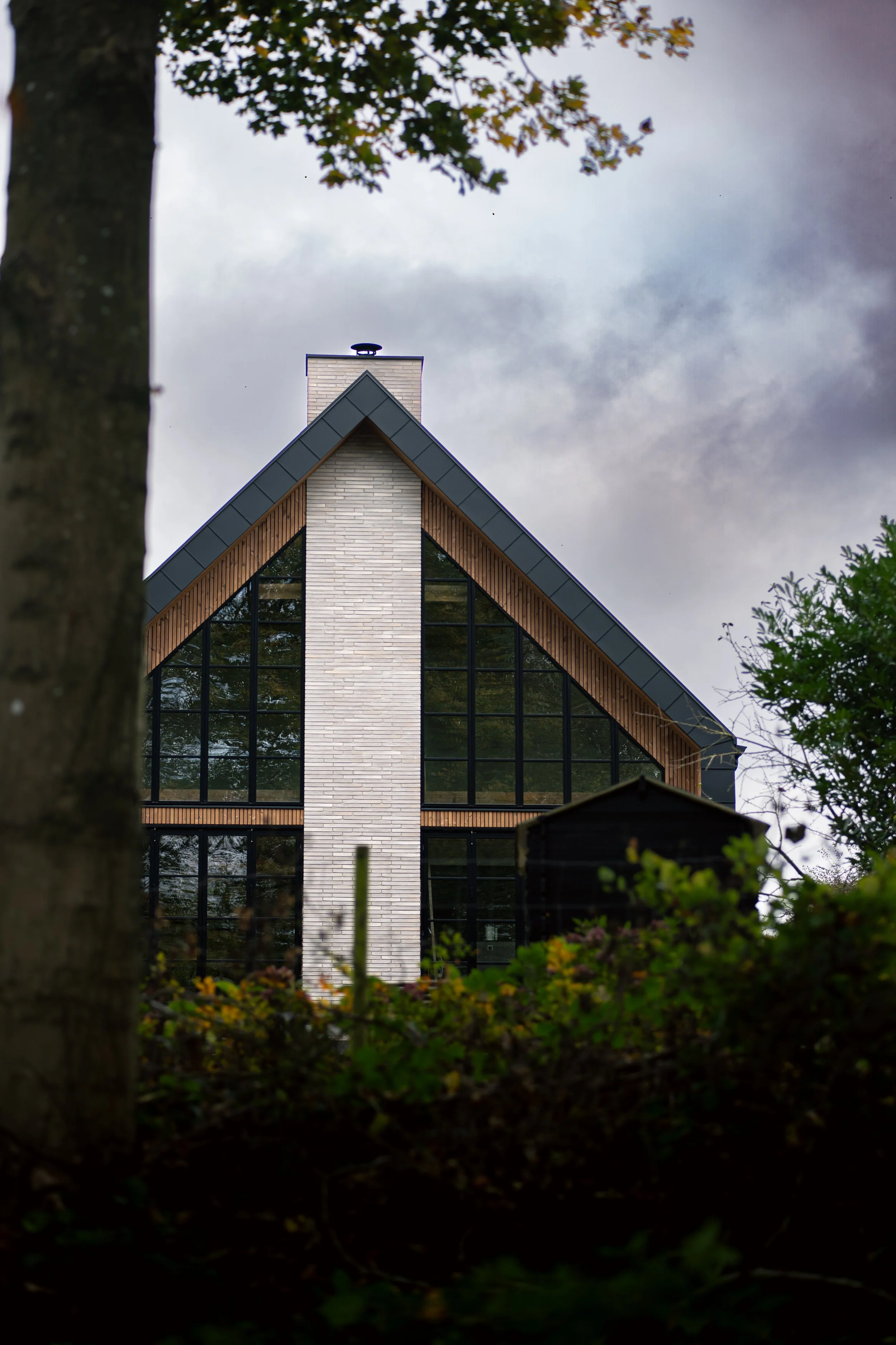 View of a modern house with large glass windows, a triangular roof, a chimney, surrounded by trees, with a cloudy sky in the background.