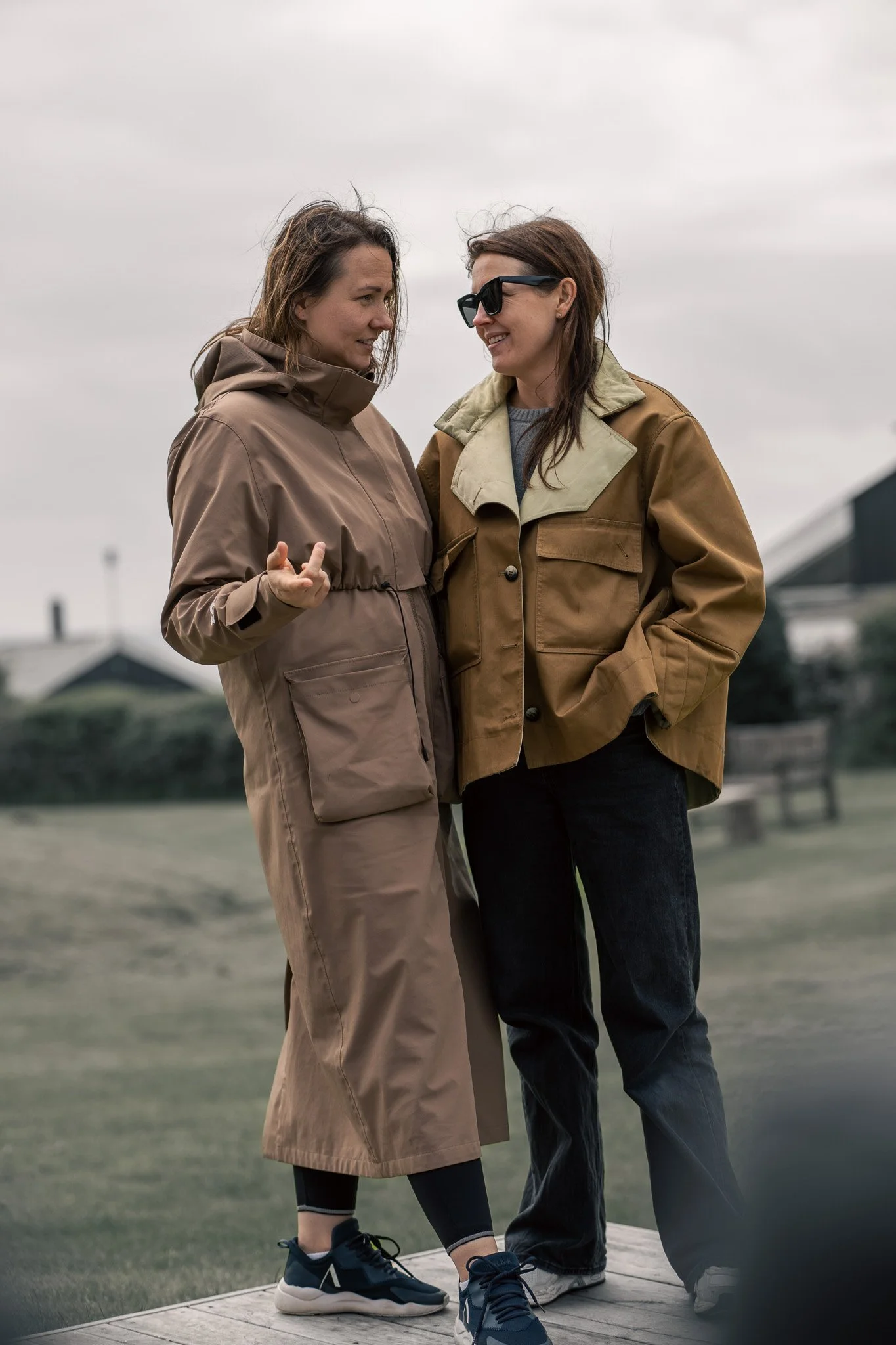 Two women are standing outside on a cloudy day, smiling and talking to each other. One woman is wearing a beige jacket and pants, and the other is wearing a brown jacket and black pants. They seem to be enjoying a casual conversation.