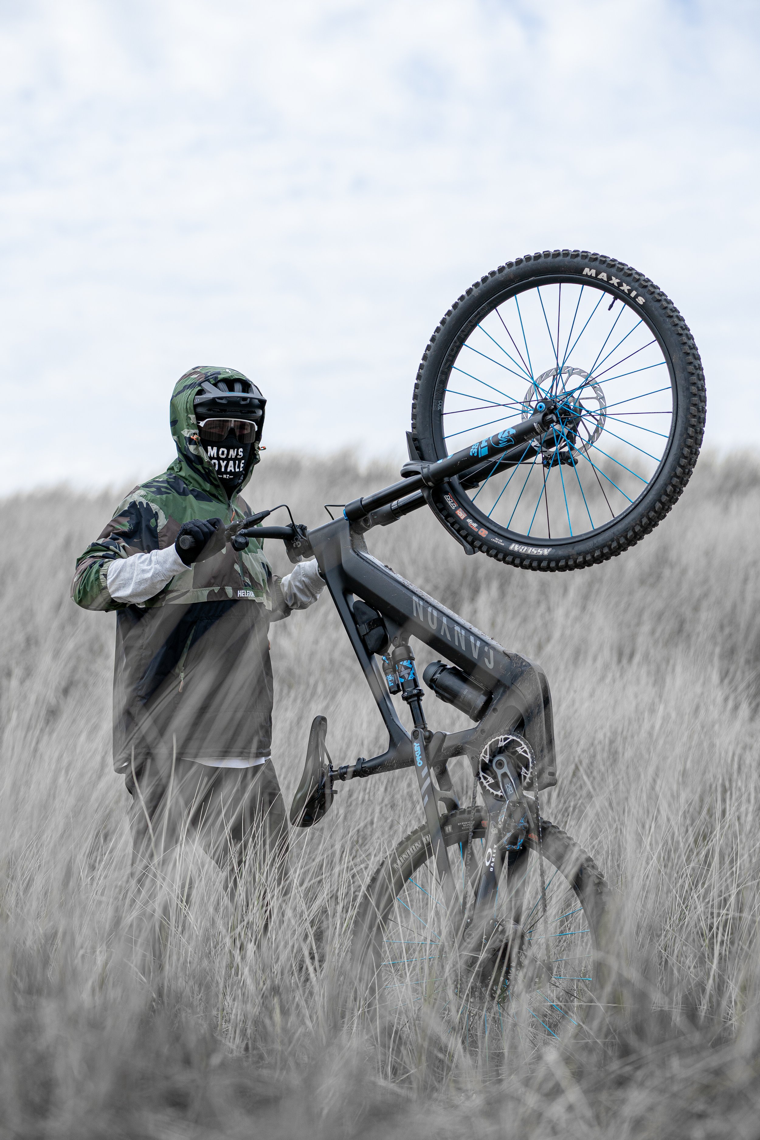 Person in mountain biking gear lifting a black mountain bike with blue accents in a field of tall grass.