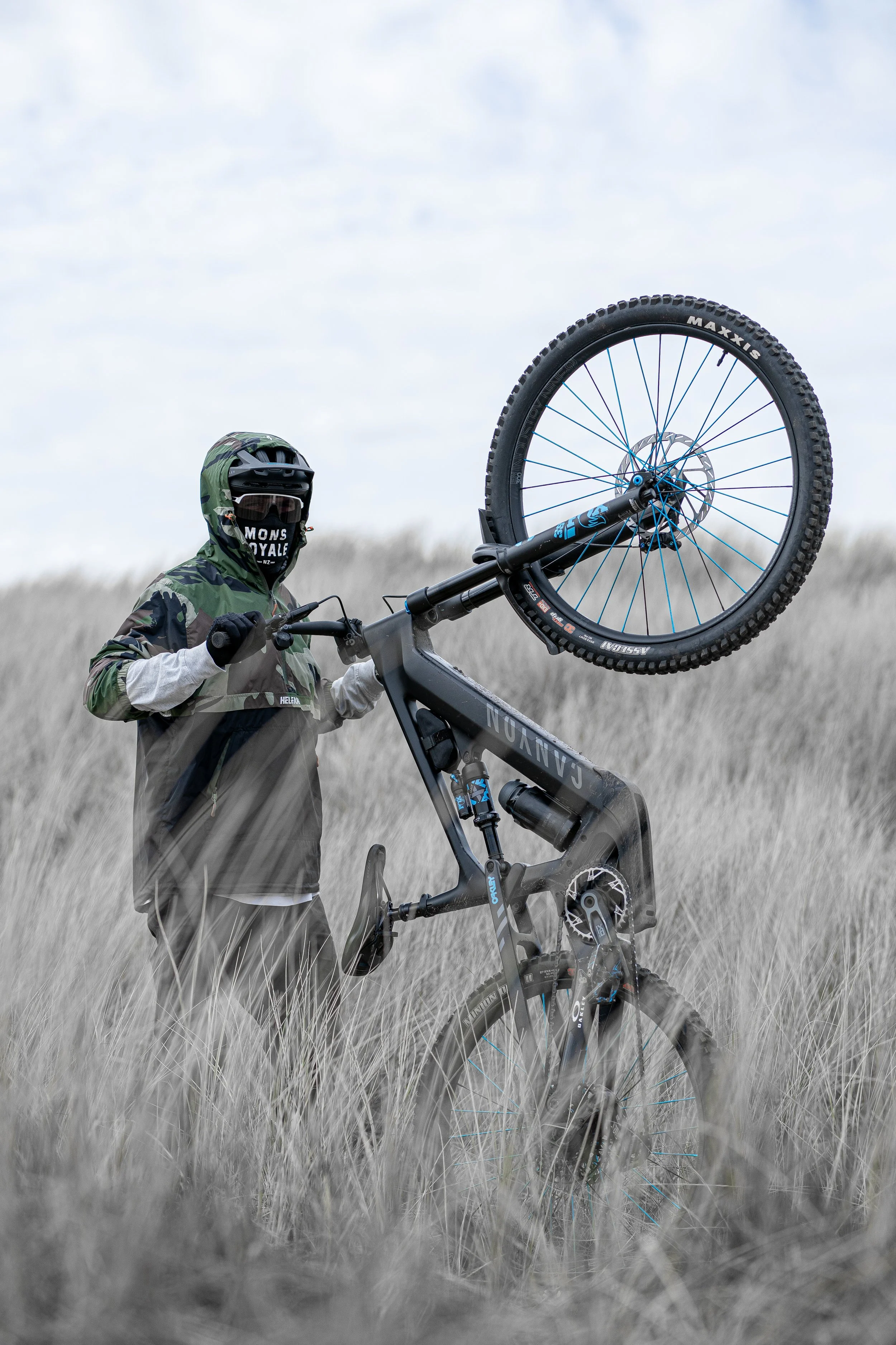 A cinematic outdoor action photo of a mountain biker lifting the front wheel in tall grass under an overcast sky, captured with a minimalist, moody composition and soft natural light.