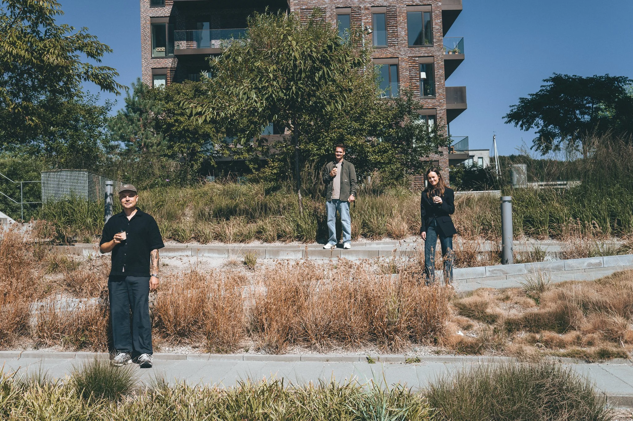 Three people standing outdoors in front of a modern apartment building, holding drinks, with trees and dry grass in the background.
