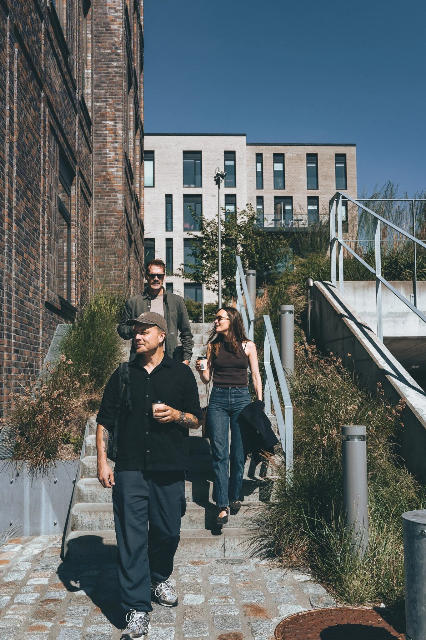Four people walking down outdoor stairs beside brick and modern buildings. The man in front has tattoos and is holding a coffee cup. The others are chatting and also holding drinks. The setting is urban with plants and blue sky.