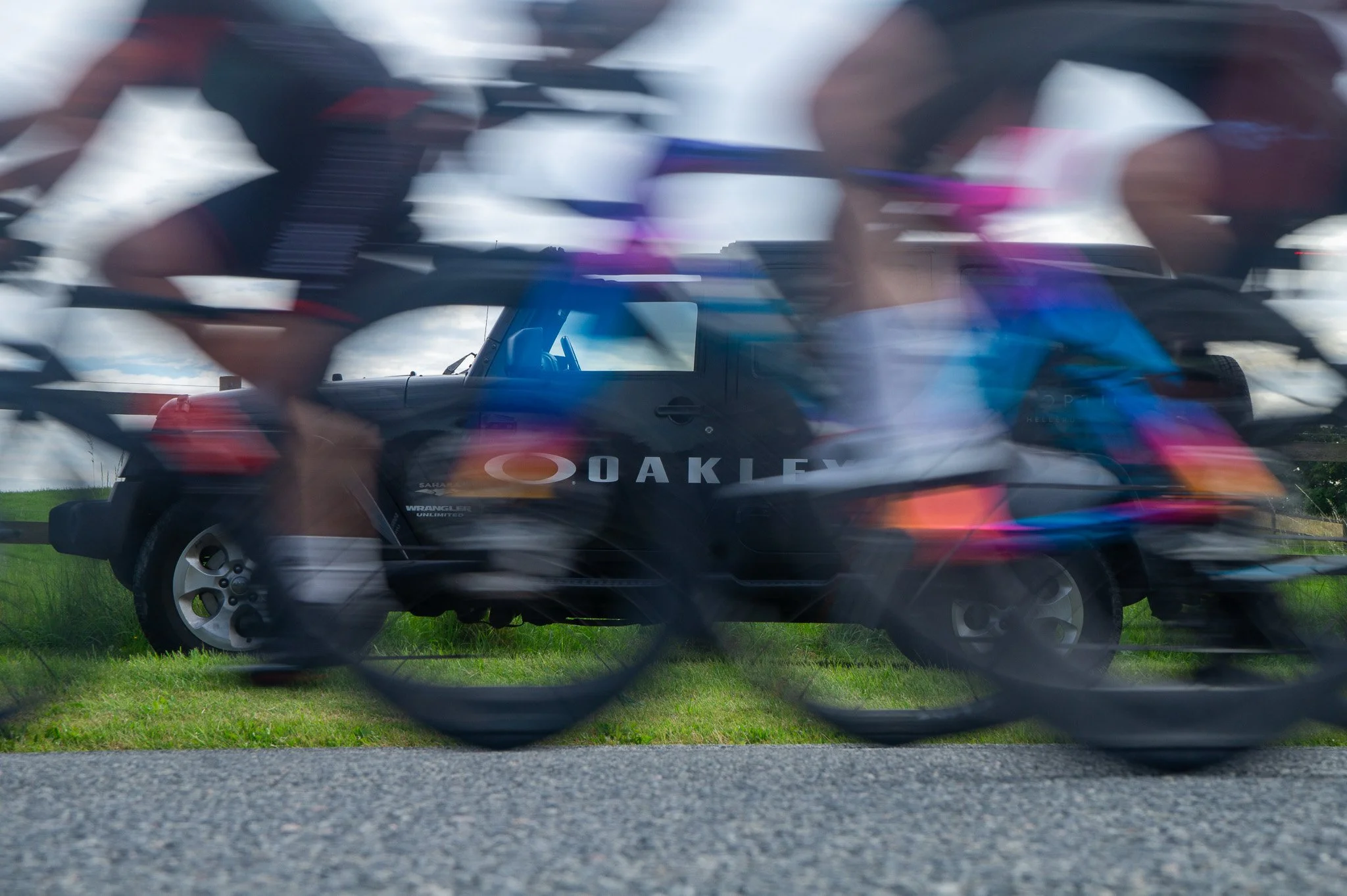 Blurry photo of cyclists riding past a black vehicle with Oakley logo on it, seen through a chain-link fence.