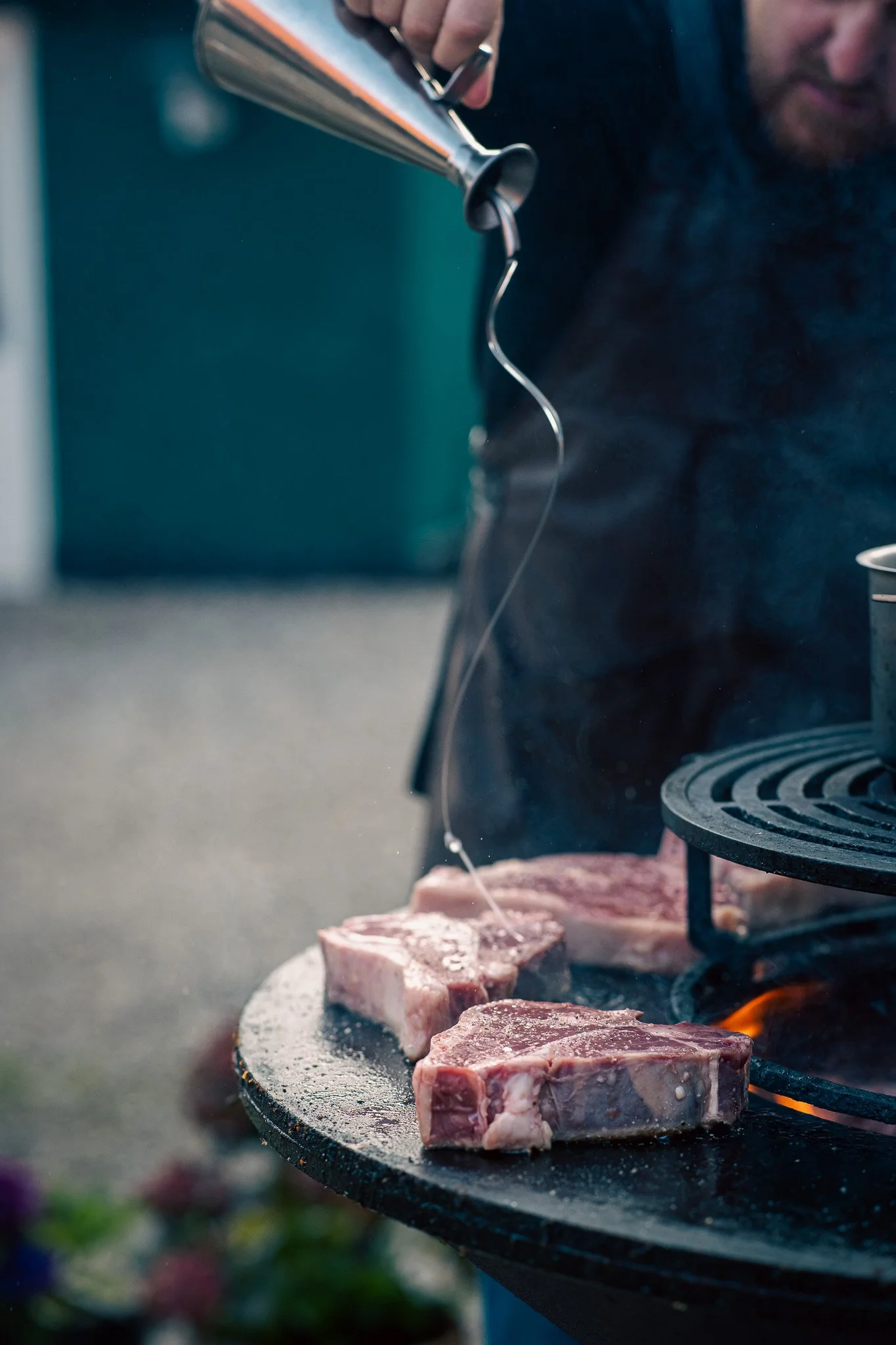 Close-up of two raw, seasoned steak cuts on a grill with a person pouring oil or marinade over them.