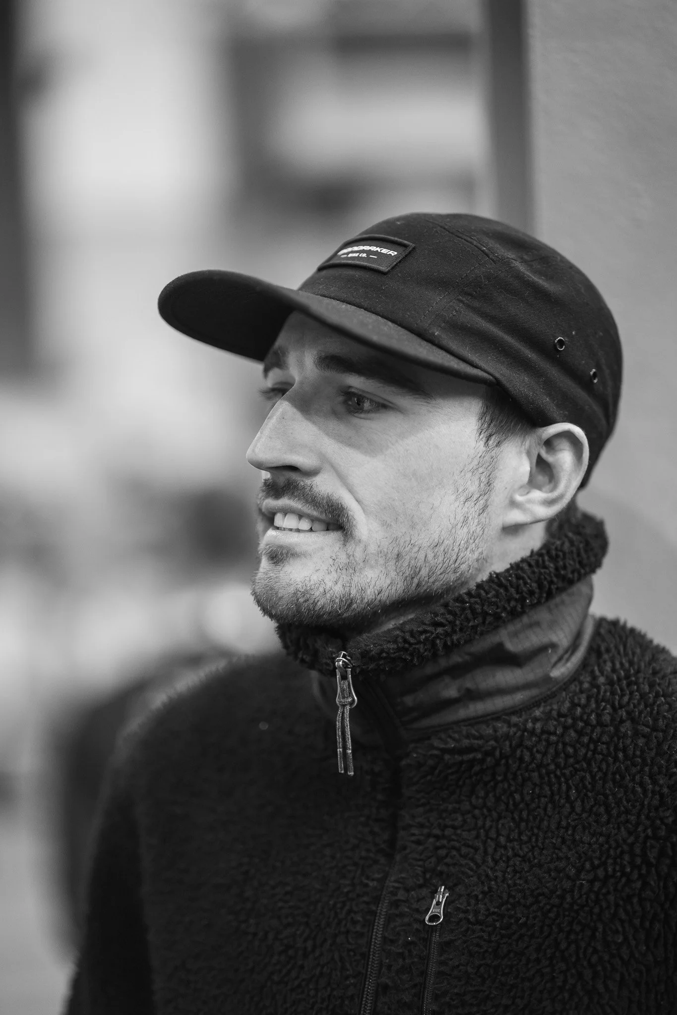 A young man in a black cap and fleece jacket, smiling in black and white.