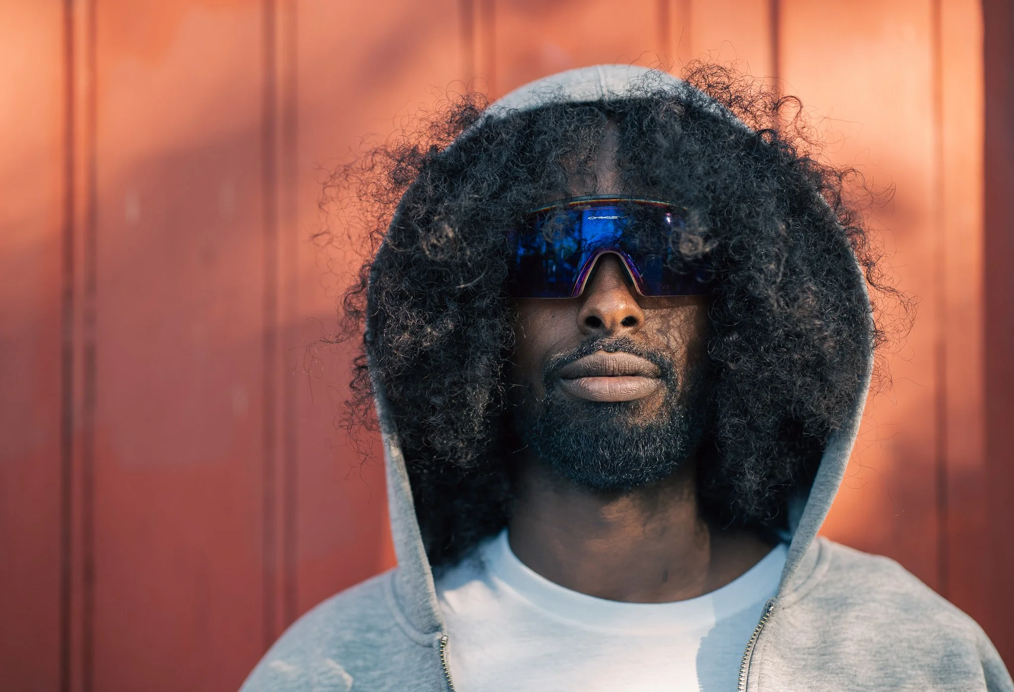 A man wearing reflective sunglasses, a grey hoodie with the hood up, and a white shirt, standing in front of a reddish-brown wall.
