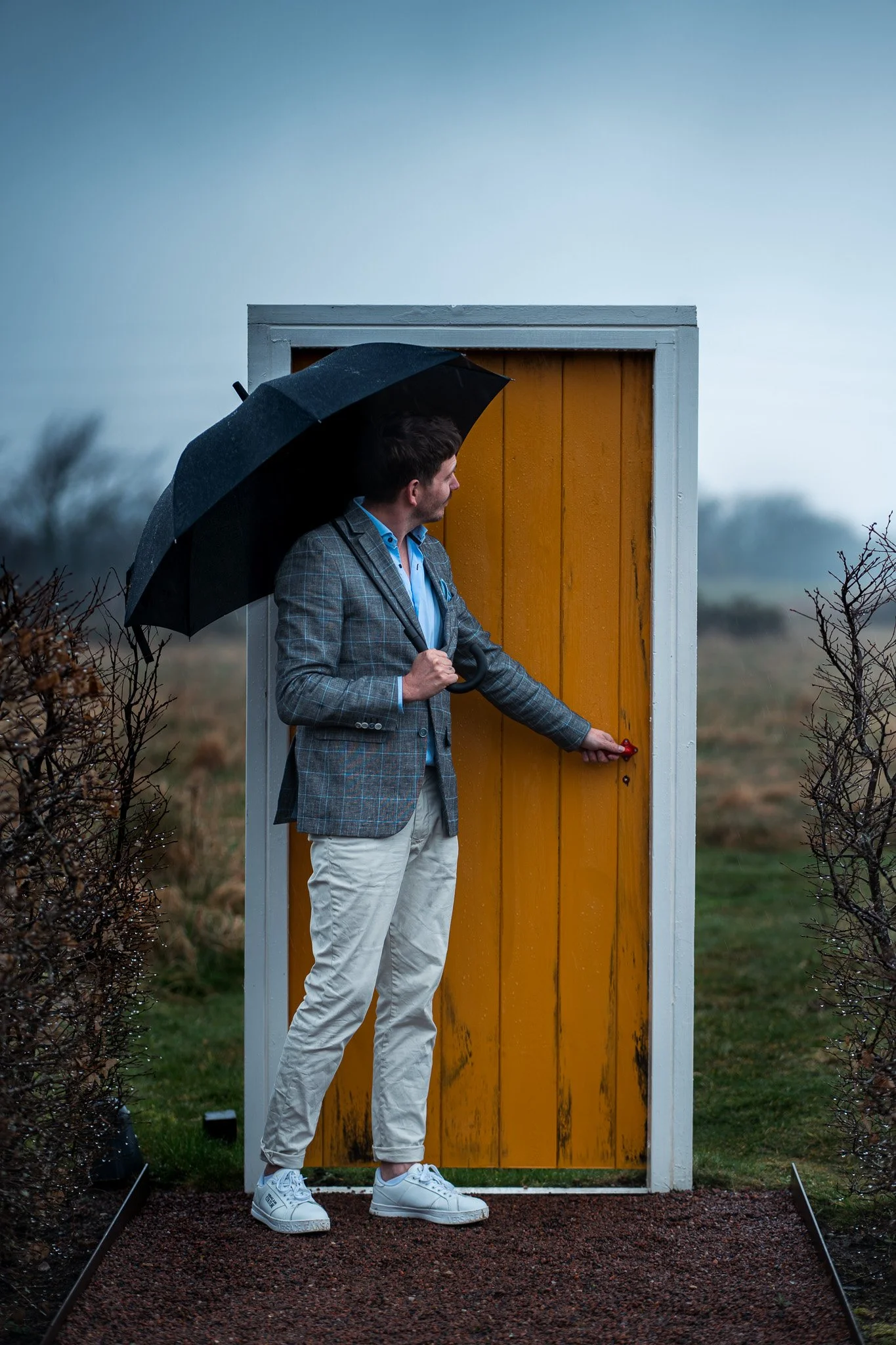 A man in a gray plaid blazer and beige pants stands in front of a yellow door, holding a black umbrella, and is about to open the door with his hand during rainy weather.