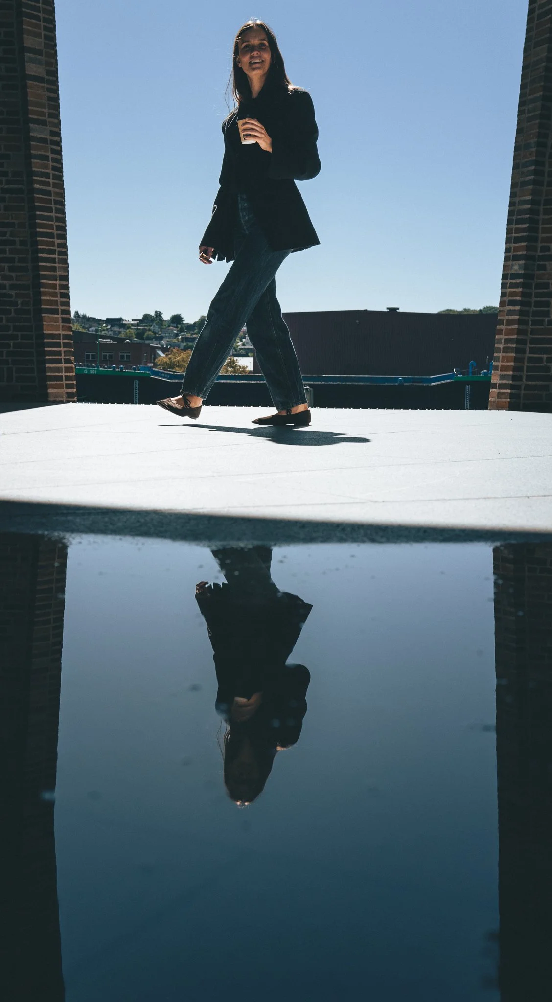 A woman standing on an outdoor balcony, holding a coffee cup, with her reflection visible in a puddle below. She is wearing a black blazer, dark pants, and flats. The sky is clear and blue.