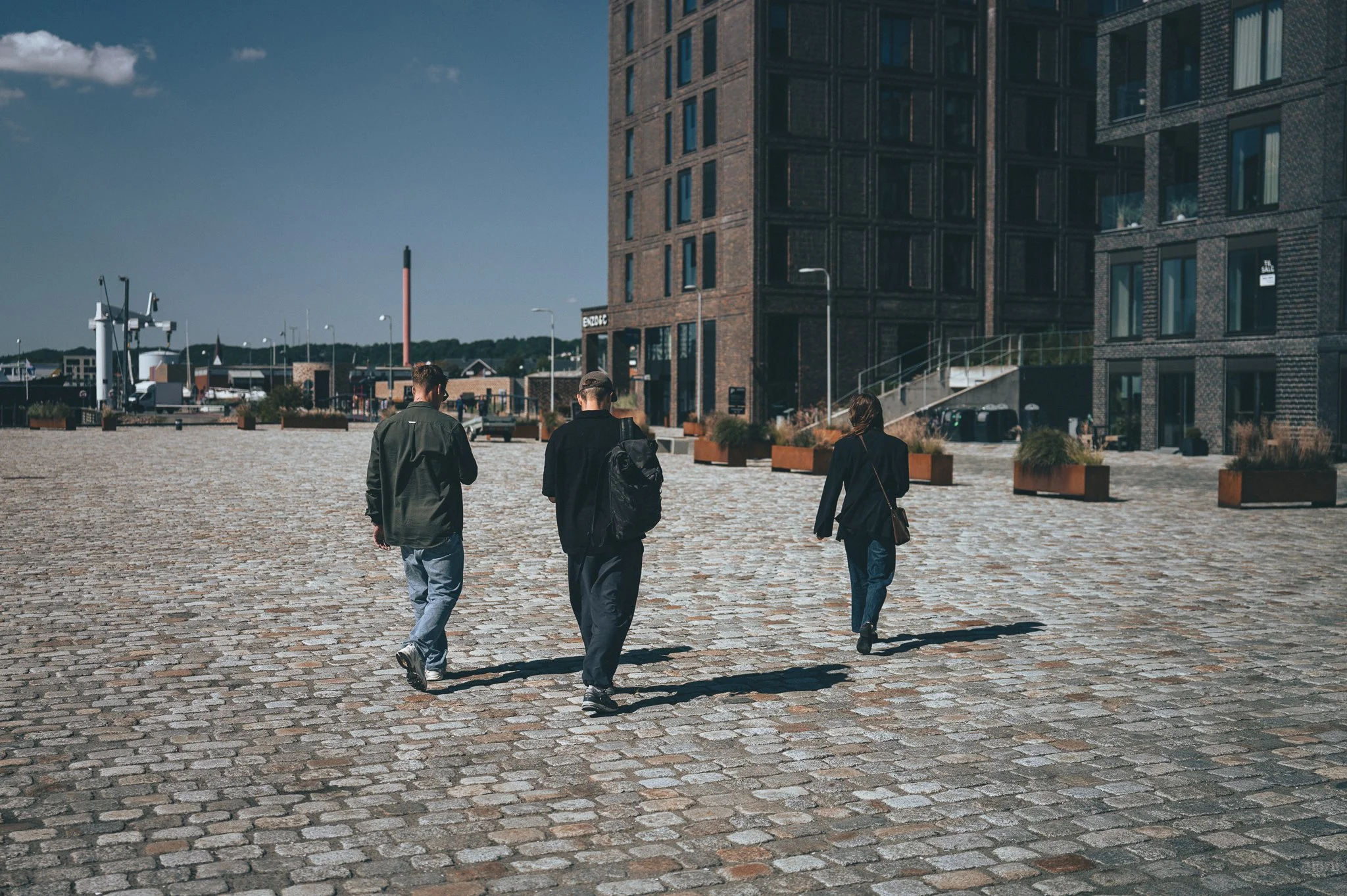 Three people walking on a cobblestone path near modern buildings during daytime.