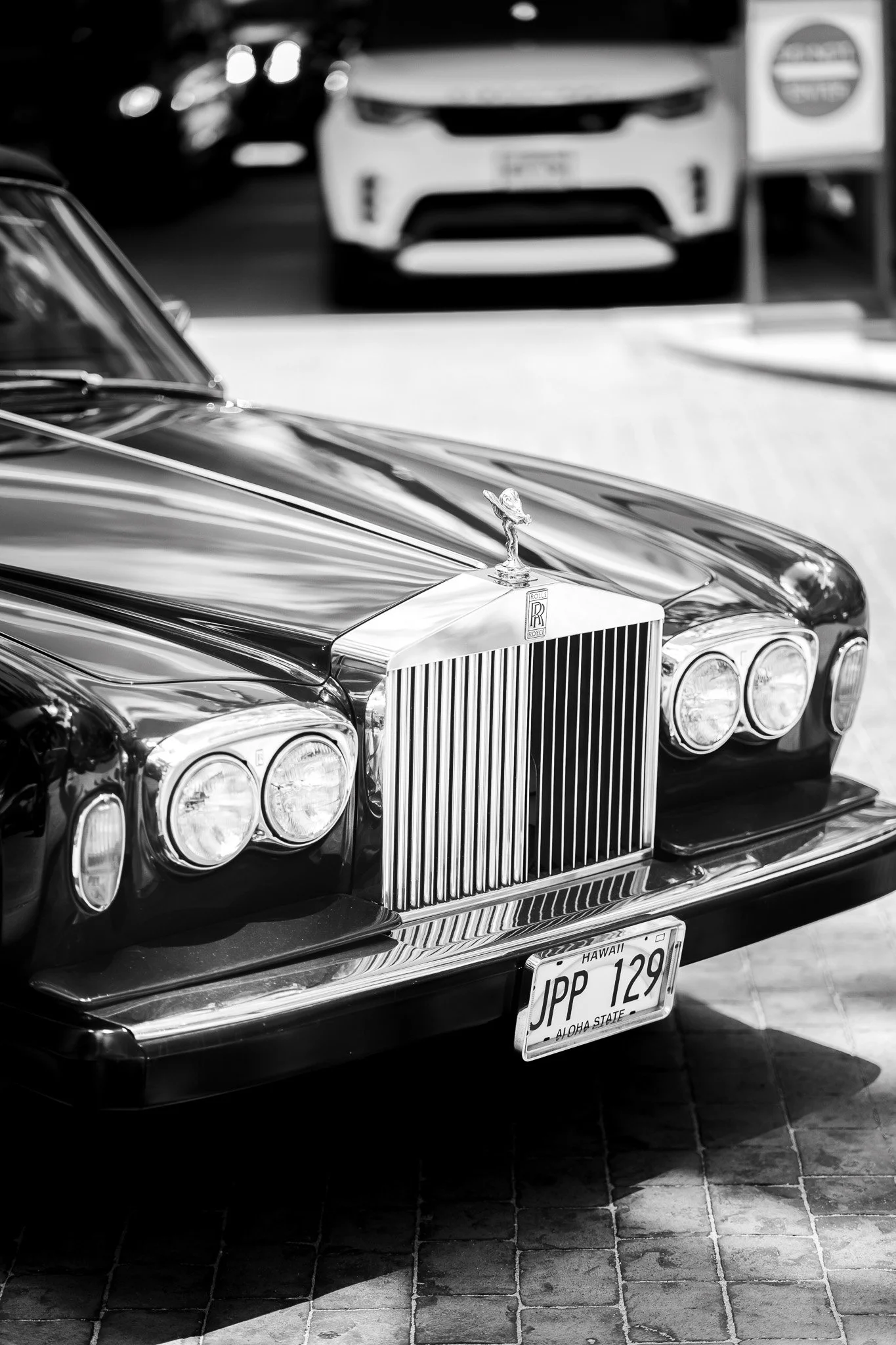Close-up of a classic black Rolls Royce car with a shiny hood ornament, parked on a brick pavement. In the background, there is a white car and a Honolulu license plate reading "JPP 129," indicating Hawaii, Aloha State.