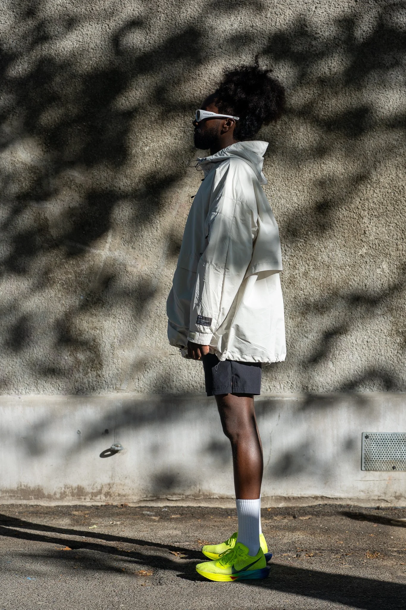 A man standing outdoors against a textured wall, dressed in athletic wear including a white windbreaker, black shorts, white socks, and bright yellow running shoes, wearing sunglasses, with shadowed tree branches overhead.