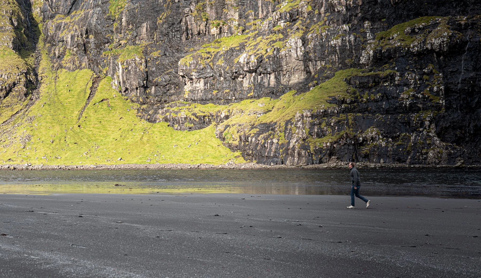 A person walking on a dark sandy beach with a large rocky cliff covered in green moss in the background.