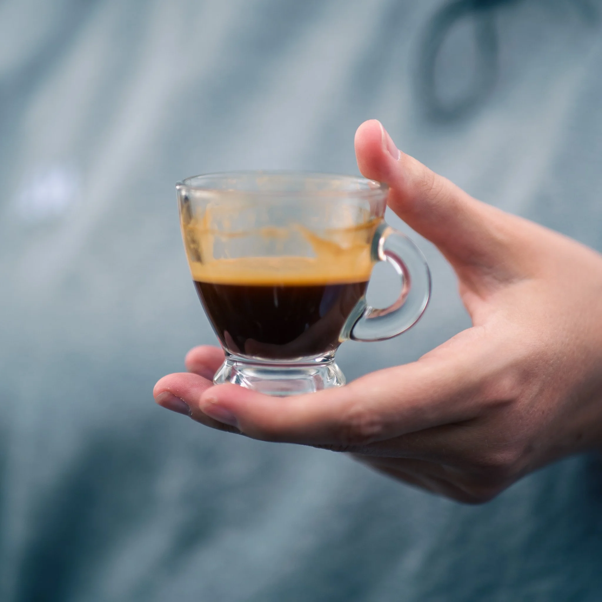 A person holding a small glass cup filled with black coffee, with a splash of coffee visible on the inside of the glass.