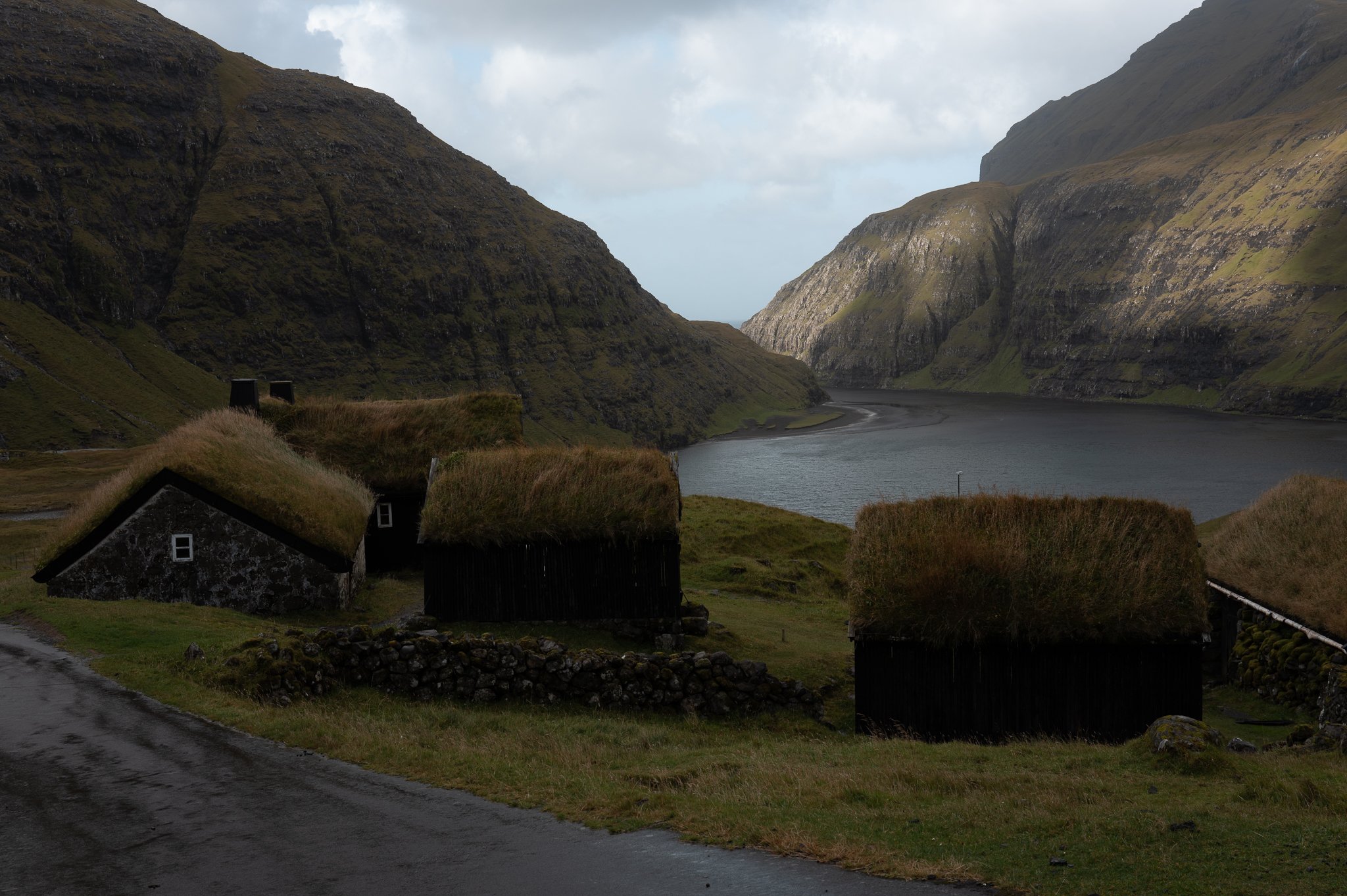 A scenic view of a narrow river surrounded by steep, grassy mountains. Several small houses with grass roofs are in the foreground near a winding road, with cloudy skies overhead.