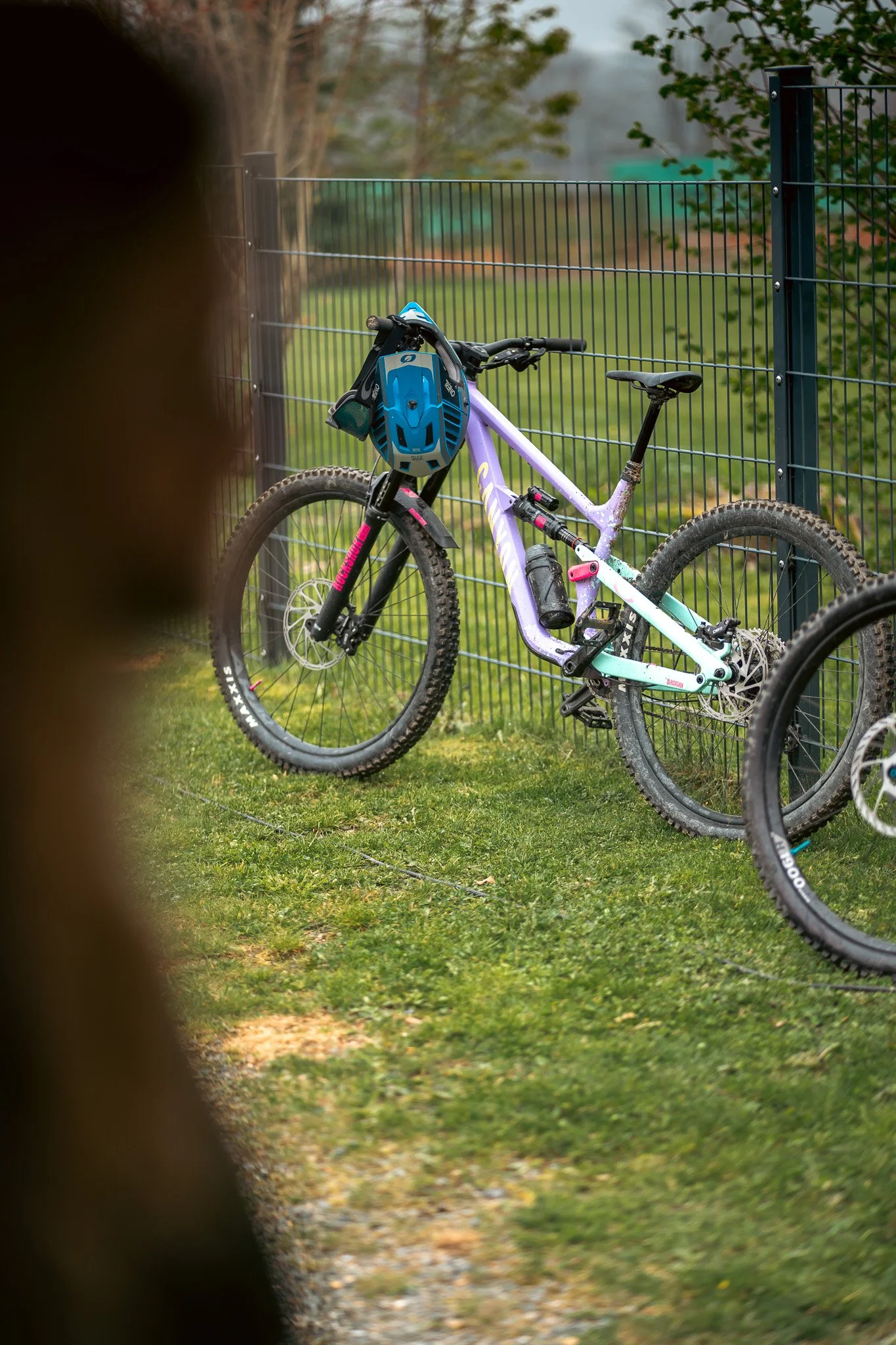 A mountain bike with a purple and light blue frame, a blue helmet hanging on the handlebar, and a water bottle mounted on the frame, leaning against a black wire fence on a grassy outdoor area.