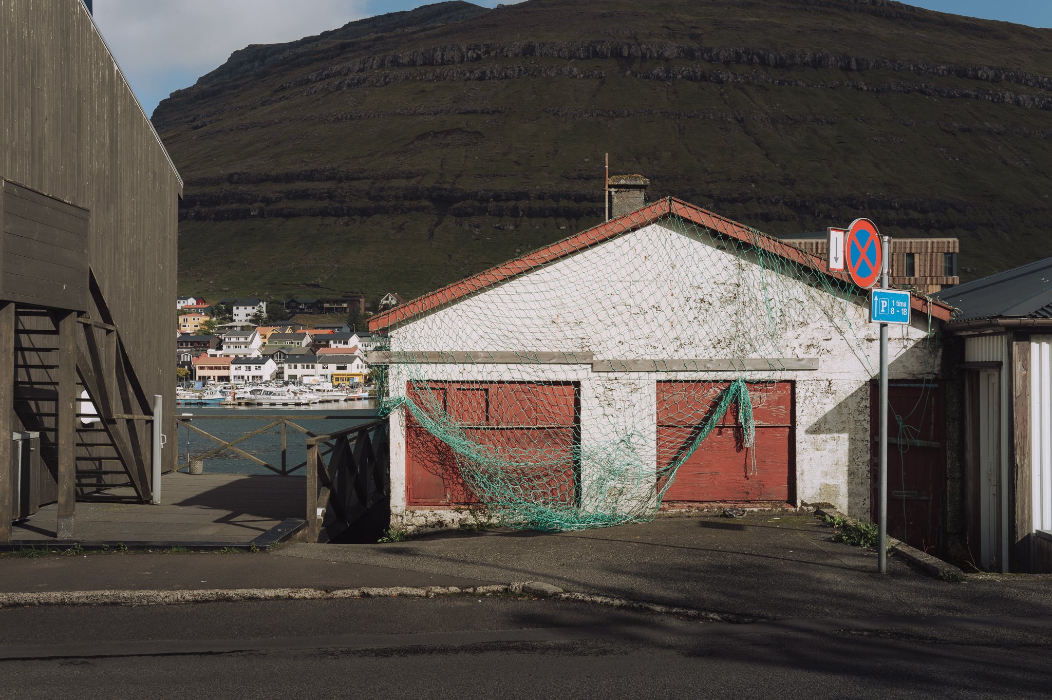 A small coastal town with colorful houses and boats docked in the harbor. In the foreground, there are wooden and concrete structures, including a building with a red roof and a view of a mountain in the background. Signs indicating parking regulatio