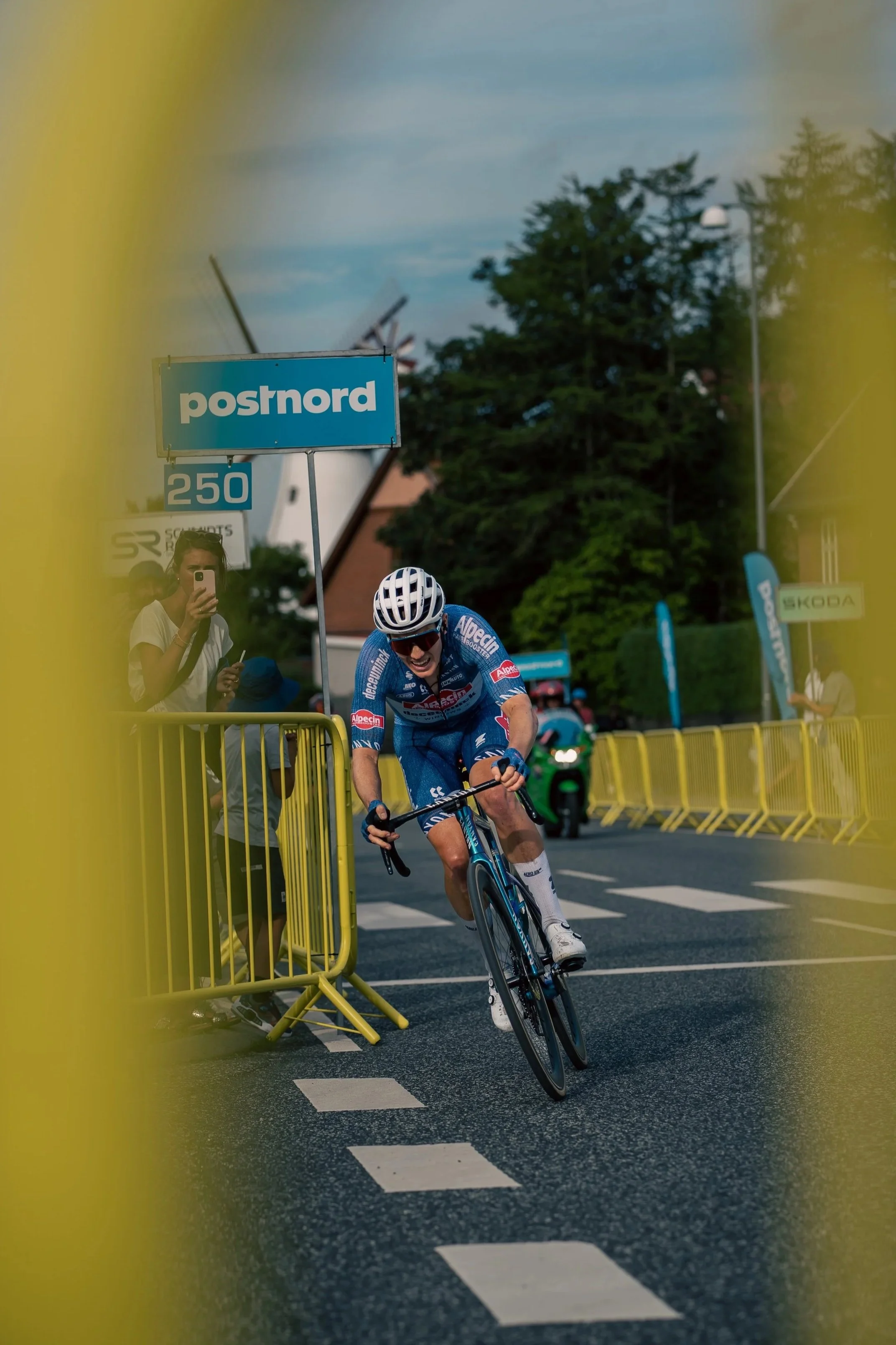 Professional road cyclist sprinting through a race finish area, captured in motion with crowd barriers, branding, and intense competitive energy.