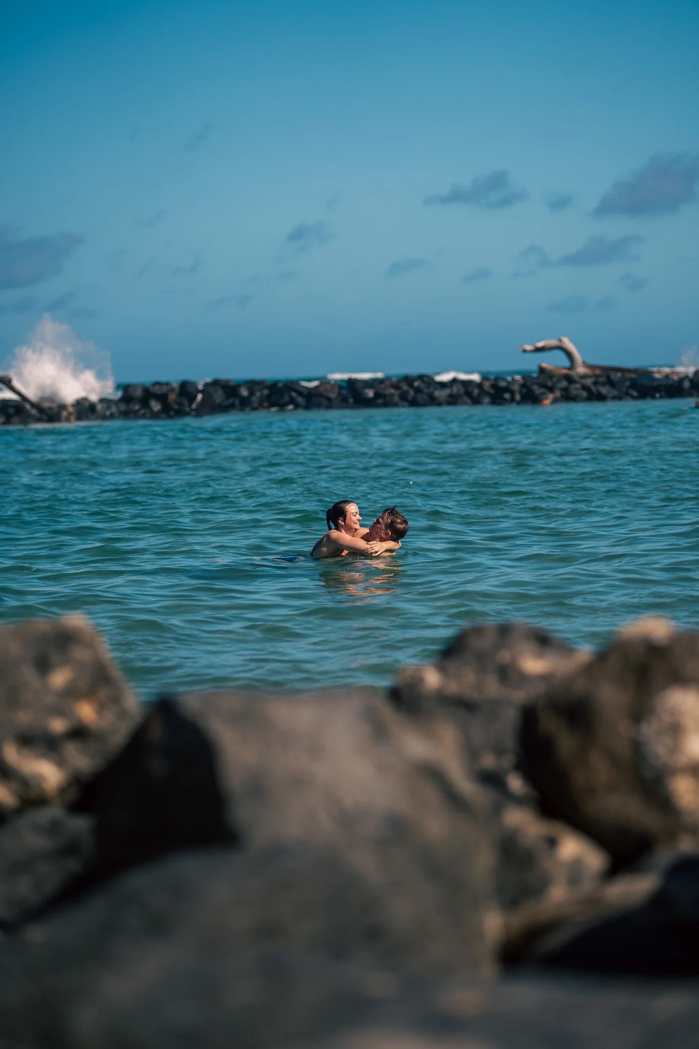 Couple swimming and hugging in ocean near rocky shoreline with breakwater and blue sky in the background.