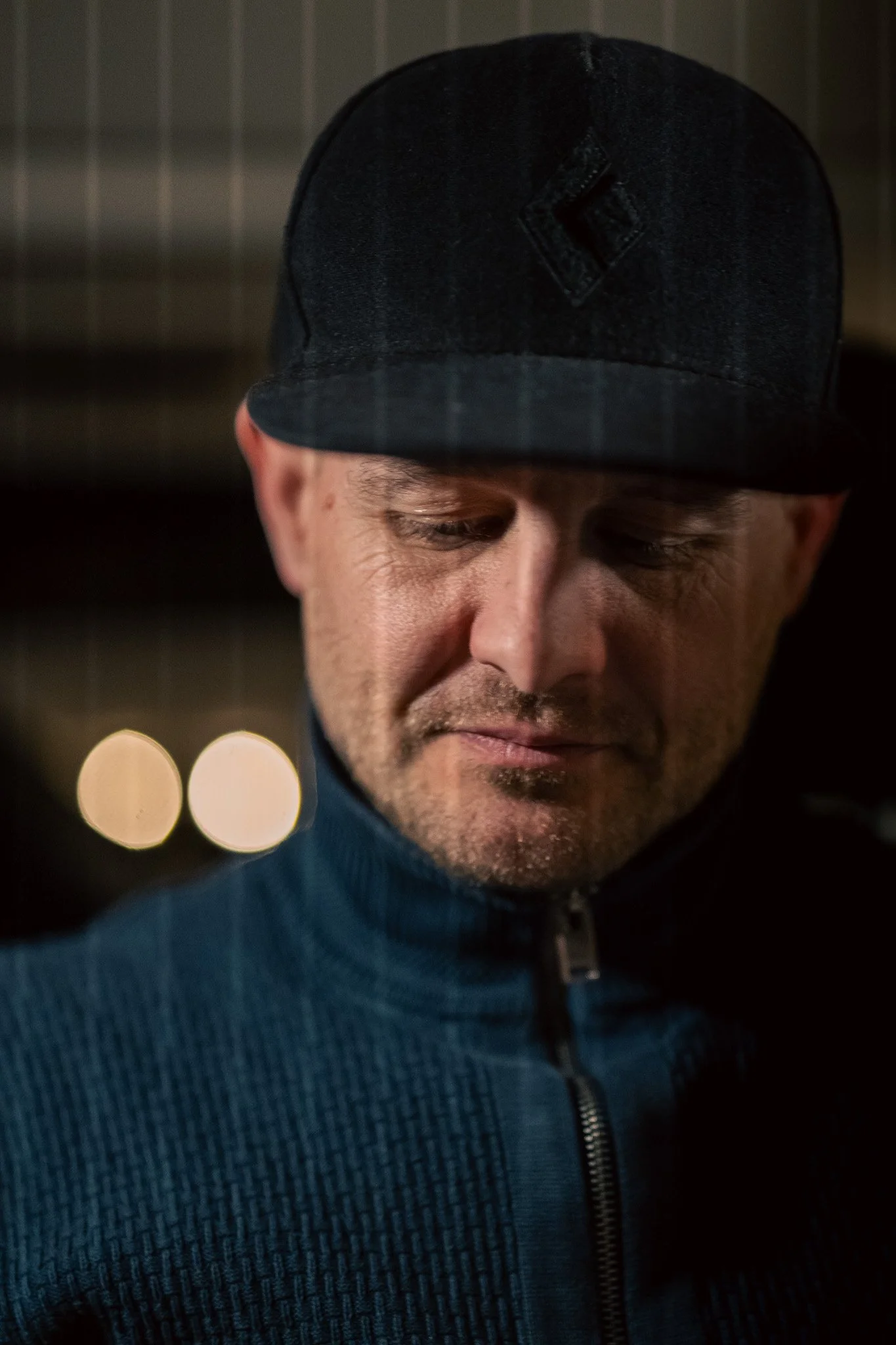 Close-up of a man wearing a black cap and blue jacket, looking down with a contemplative expression.