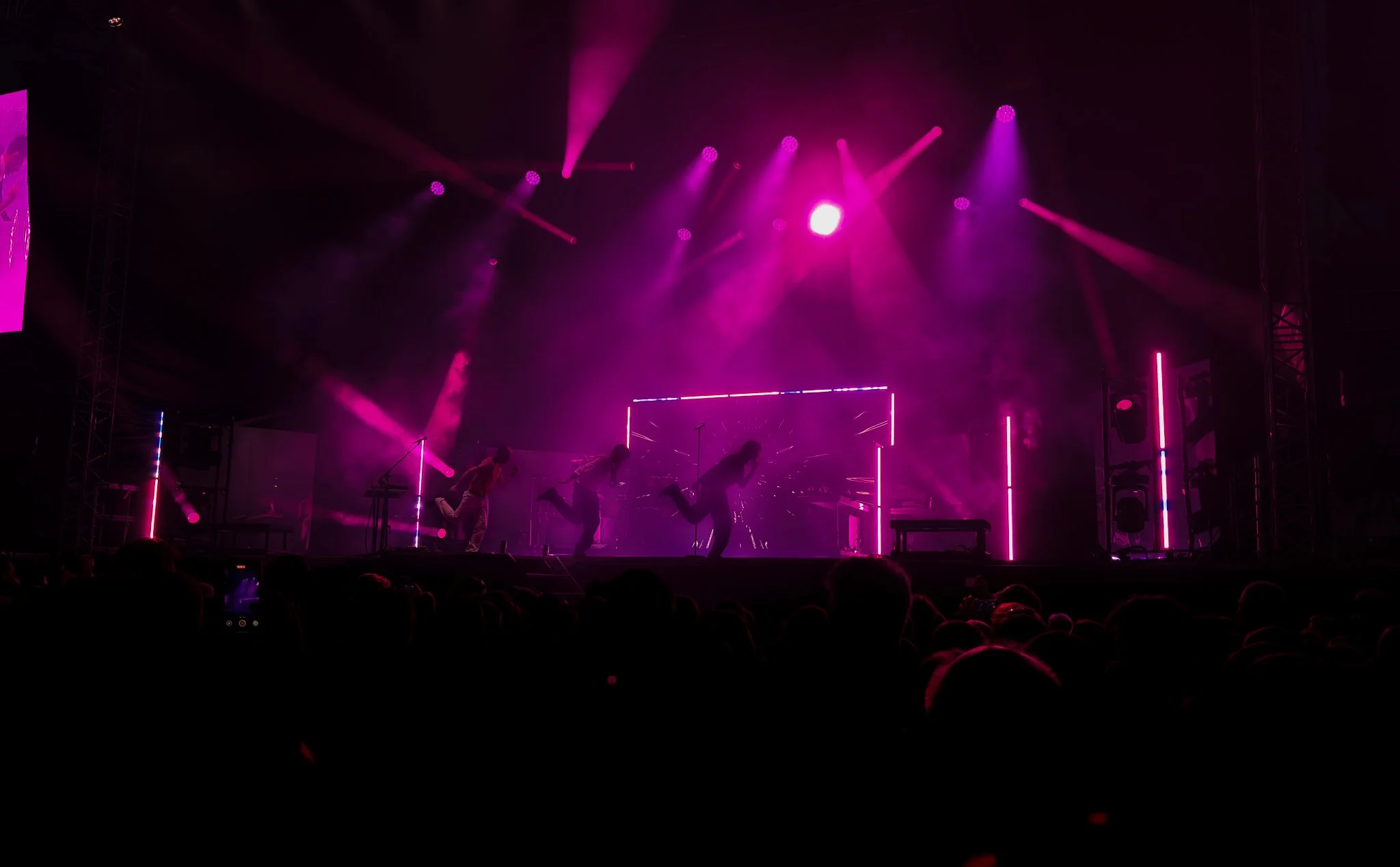 A live concert with a stage illuminated in pink and purple lighting. Three performers are dancing or jumping, and there are bright visual effects on the screen behind them. The audience is visible in silhouette in the foreground.