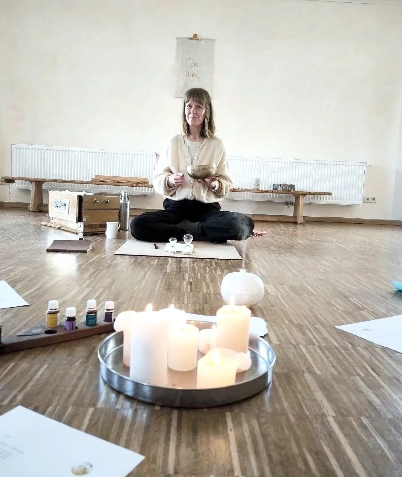 Eine Frau sitzt auf dem Boden in einem Raum, hält eine Klangschale und hat verschiedene Gegenstände um sich herum, inklusive Kerzen und ätherischer Öle, die für eine Klangmassage oder Meditation verwendet werden.