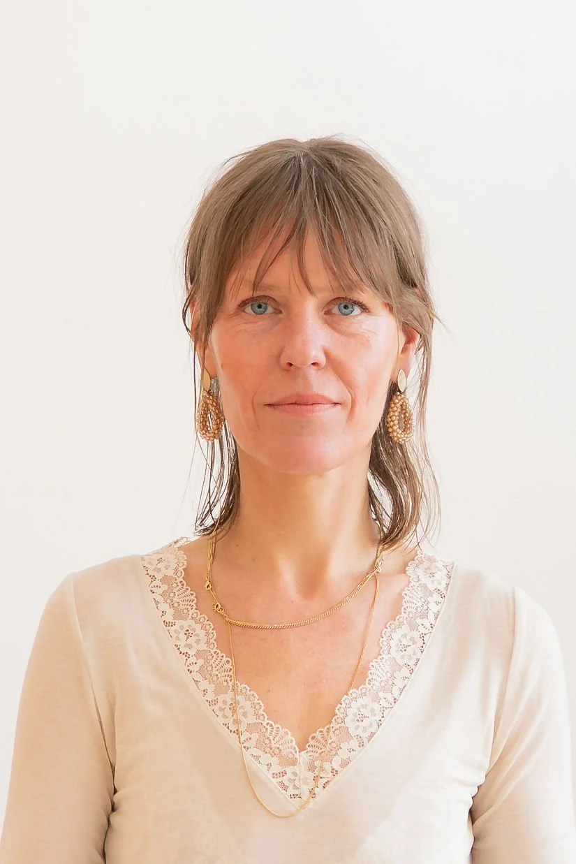 Porträt einer Frau mit braunen glatten Haaren, blauen Augen, die eine cremefarbene Bluse mit Spitzenverzierungen trägt, sowie goldene Ohrringe und Halsketten, vor hellem Hintergrund.