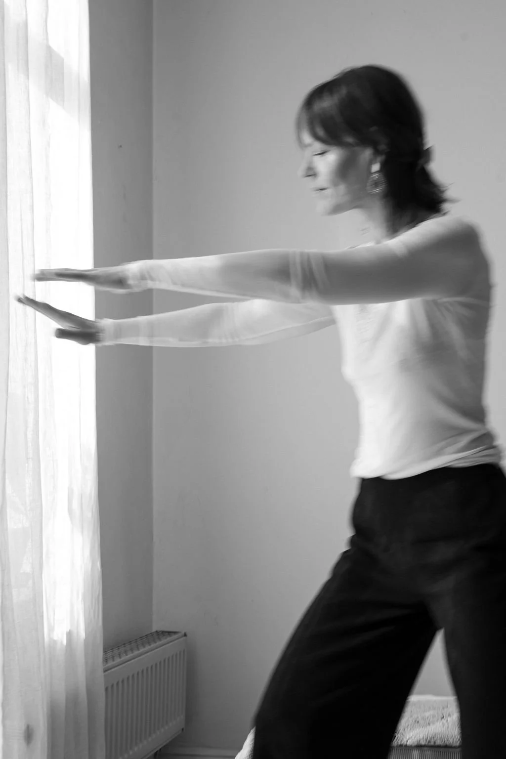 Frau in schwarzer Hose und weißem Top macht eine Dehnübung im Raum, blickt lachend nach rechts, steht neben einem Fenster mit halb transparenten weißen Vorhängen.